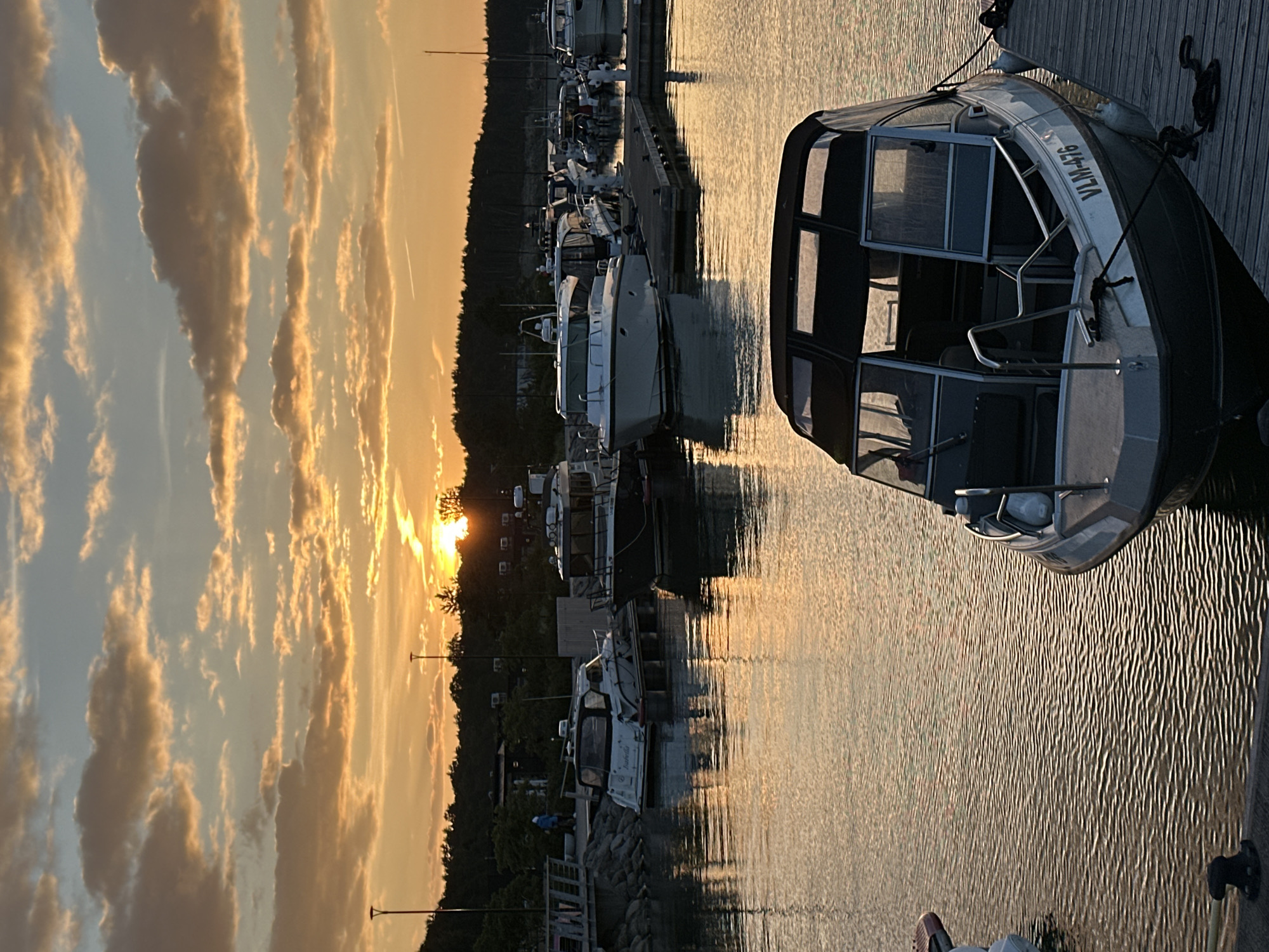 The image depicts a serene marina scene at sunset in Tallinn, Estonia. The sky is painted with warm hues of orange and yellow, with scattered clouds catching the last rays of sunlight. The water in the marina reflects the vibrant colors of the sky, creating a shimmering surface.
In the foreground, a sleek motorboat is docked, its dark canopy contrasting with the bright reflections on the water. The boat has the registration number "VLM-476" visible on its hull. The boat is secured to a wooden dock with dark ropes and fenders.
Further into the marina, several other boats are moored. One white boat with a dark canopy is positioned to the left, with the name "Isabella" visible on its stern. Other boats, varying in size and color, are lined up along the docks.
The background reveals a densely wooded shoreline, with trees silhouetted against the twilight sky. A few structures, possibly part of the marina or coastal buildings, are visible amongst the trees. The overall atmosphere is tranquil and picturesque, capturing the beauty of a summer evening in a coastal city.