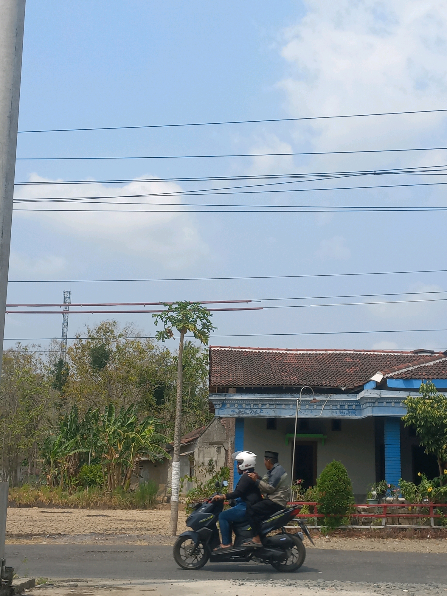 In Baturetno, Indonesia, on a clear, sunny day, two individuals are riding a black scooter. The person in front, wearing a white helmet and a black long-sleeved shirt, is driving. The passenger behind, wearing a black cap and a light-colored jacket, has their right arm around the driver. They are traveling on a paved road. To the right of the road is a blue and white house with a red tile roof, surrounded by greenery, including a small tree and potted plants. Beyond the house and to the left, there are more trees, some of which appear to be banana trees, and a distant cell tower. Overhead, multiple power lines stretch across the sky, and a tall, thin pole with white wrapping is visible on the right side of the image.