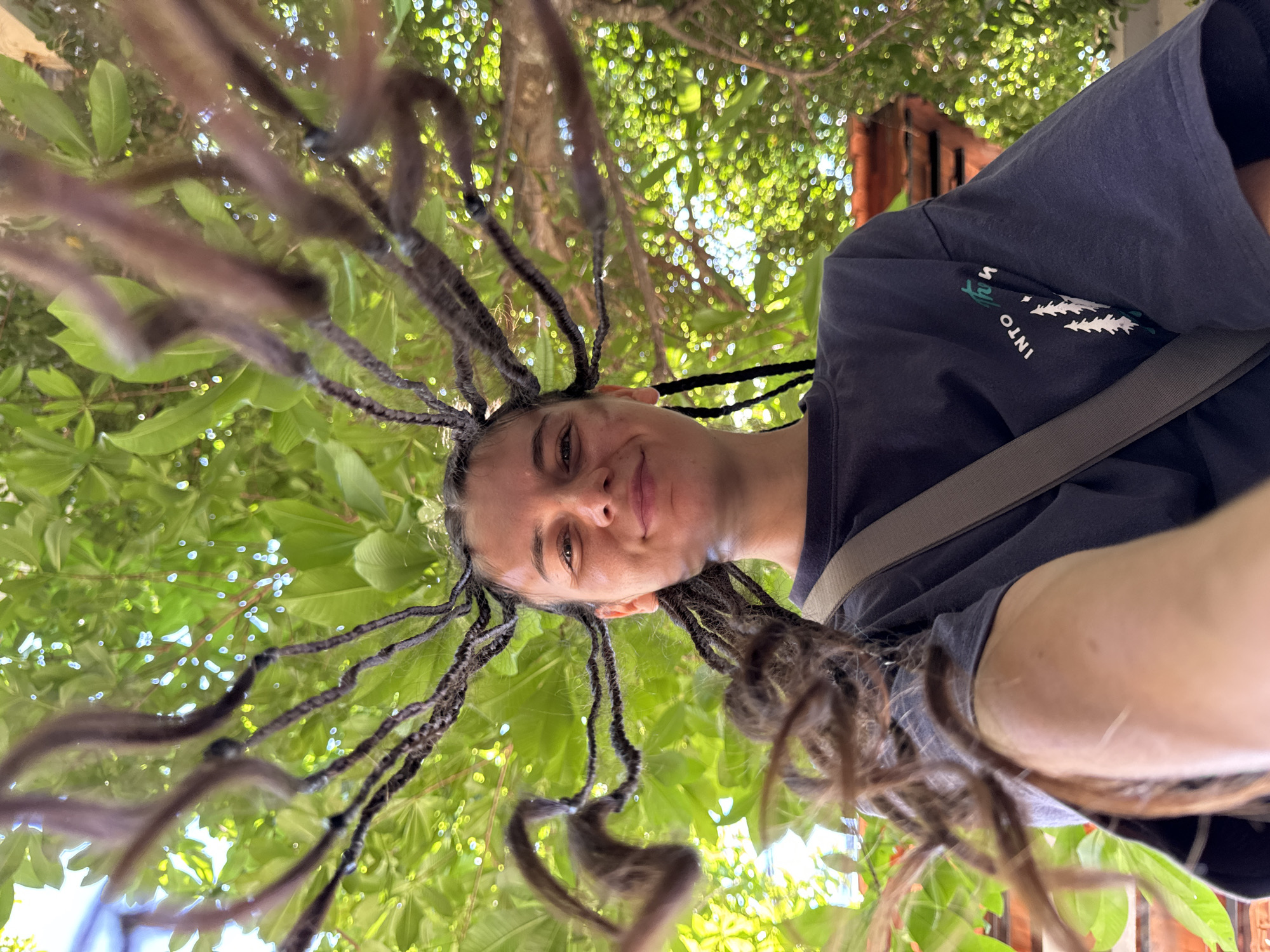 A young woman with dreadlocks is taking a selfie outdoors, looking up at the camera with a slight smile. Her dreadlocks fan out around her head, creating a dramatic effect against the bright green foliage of trees above. She is wearing a dark t-shirt with a graphic print that appears to be trees and text. A grey strap is visible across her chest, suggesting she may be wearing a bag. The background is filled with lush green leaves and sunlight filtering through, indicating a natural or outdoor setting. The image appears to have been taken from a low angle, looking up towards the woman and the trees. There are no other people or significant objects visible in the frame besides the woman and the natural environment. There is no discernible urban environment or location markers that would suggest the city of Mui Ne, Vietnam.