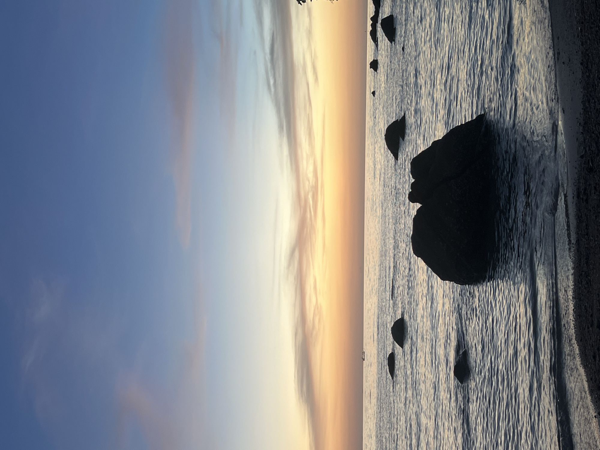 This is a vertical, full shot of a serene coastal scene at sunset. The left side of the image is dominated by a gradient sky, transitioning from a deep blue at the top to soft hues of pink, orange, and yellow near the horizon, indicative of twilight. Wispy clouds are scattered across the sky, catching the last rays of the sun.
The right side of the image depicts the ocean meeting a sandy shore. Several dark, silhouetted rocks are scattered in the shallow water, with the largest one prominently positioned in the foreground. The water has a textured appearance, with ripples reflecting the subtle colors of the sky. The shoreline, a strip of dark sand, is visible on the far right. In the distance, a small, dark boat can be seen on the calm water. The overall mood is peaceful and tranquil, likely captured during the golden hour of sunset. There is no visible text in the image.