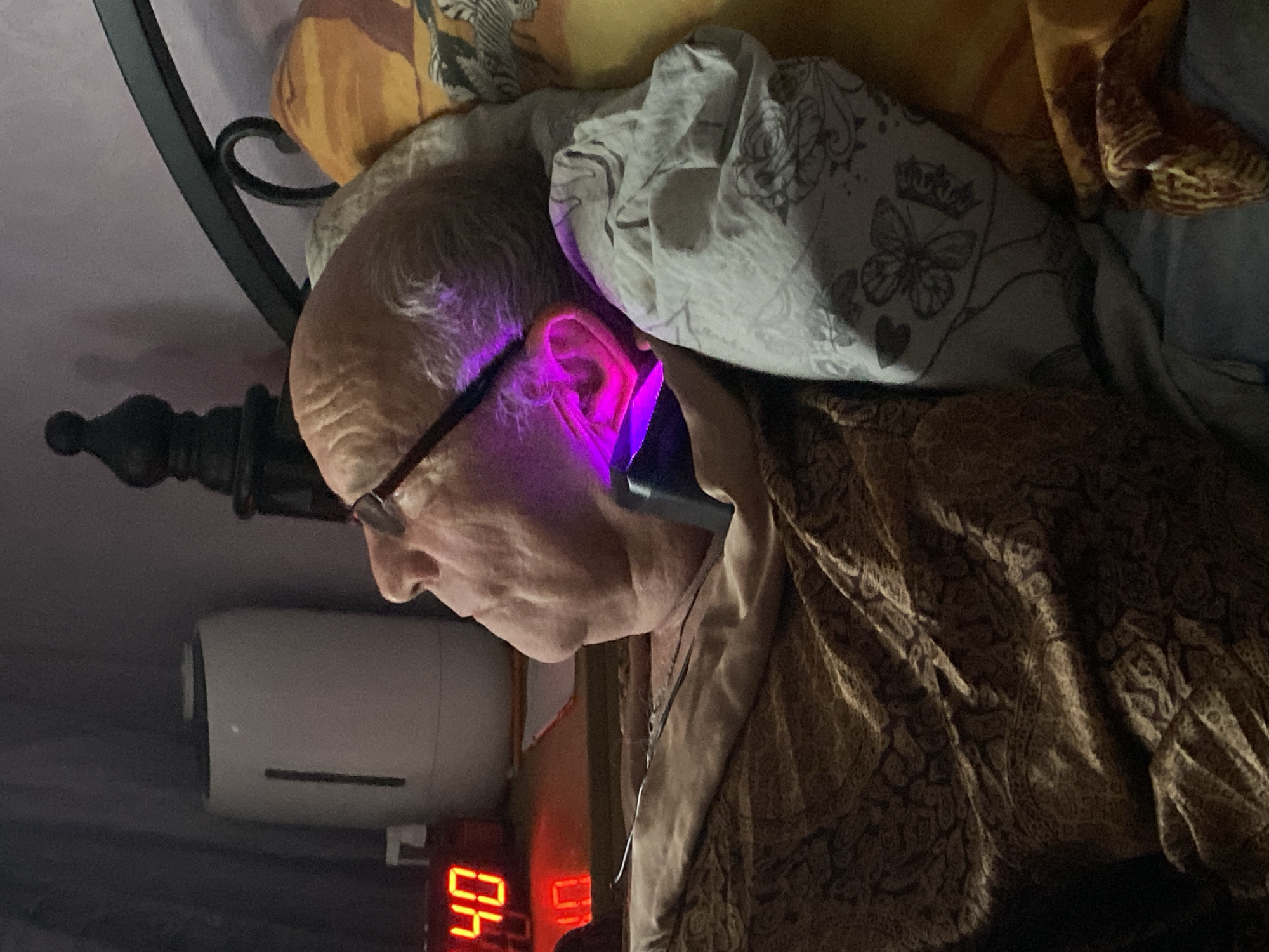 An elderly man, possibly in bed, has a bright purple light shining into his ear. He is wearing glasses and is partially covered by bedding. A digital clock displays the number "40." A humidifier is visible in the background. The location, Novokuznetsk, Russia, cannot be definitively confirmed from the image.