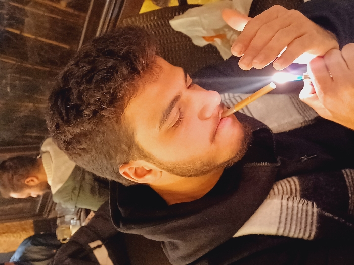 A person with dark, curly hair is shown in profile, holding a lit cigarette or joint in their mouth. Another person, partially visible in the background, appears to be sitting at a table. The main subject is wearing a dark hoodie with a white and black striped pattern underneath. A lighter is positioned near the end of the cigarette, with a flame clearly visible. The lighting suggests an indoor setting, possibly a bar or lounge.