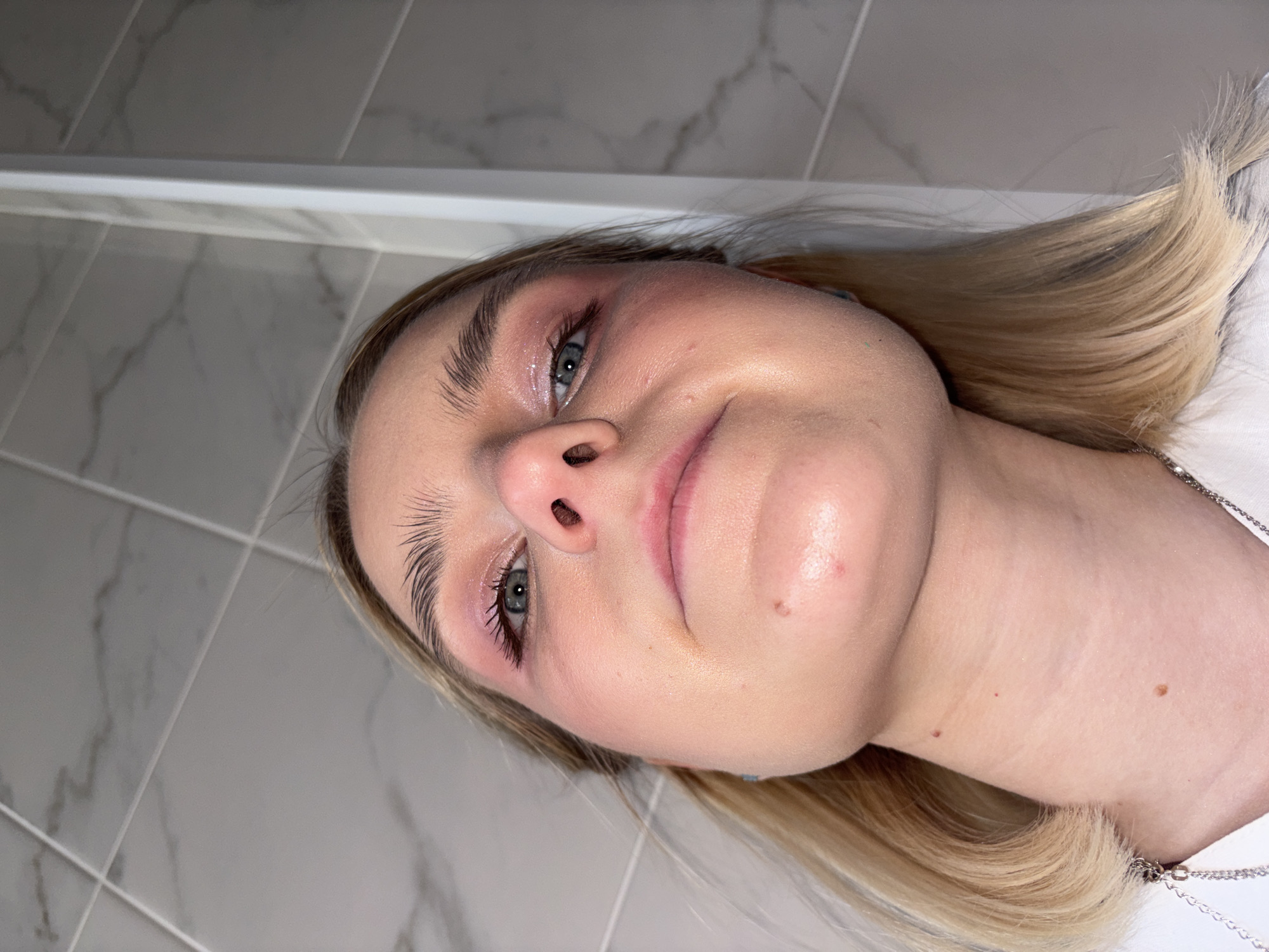 The image is a close-up portrait of a young woman, rotated 90 degrees clockwise, showing her face from a low, upward-looking perspective. She has blonde hair, fair skin, and light blue-green eyes.
The subject's face is centrally framed, exhibiting a dewy or glossy complexion. Her makeup includes groomed, dark eyebrows, pinkish eyeshadow with a shimmery highlight on the eyelids, and dark mascara on her eyelashes. There are minor blemishes and visible pores on her skin. She has a subtle, almost contemplative expression with a slight smirk on her lips. A silver-colored chain is visible around her neck, suggesting she is wearing jewelry over a white garment, possibly a t-shirt or blouse.
The background consists of light gray and white rectangular tiles, featuring a subtle marble-like pattern and horizontal grout lines. A lighter, possibly white, trim piece runs horizontally across the upper portion of the background. This tiled setting suggests an indoor location, likely a bathroom or kitchen. The lighting appears direct and artificial, creating highlights on her skin and hair.
No discernible text is visible in the image. The activity depicted is a close-up photo, likely a selfie, focusing on the subject's face and makeup. The image does not contain specific visual cues to confirm the location context of Saint Petersburg, Russia.