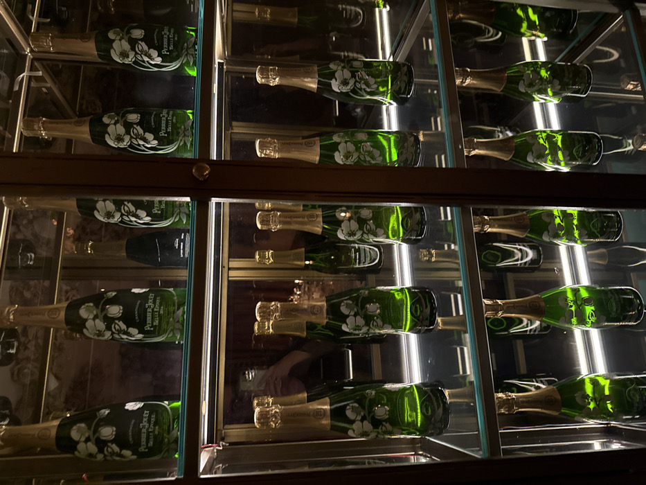 A cabinet displays multiple bottles of Perrier-Jouët Belle Epoque champagne. The bottles have a green tint and are adorned with white floral designs. They are arranged horizontally in rows within the glass-enclosed cabinet. The bottles and their reflections are prominent due to the lighting inside the cabinet.