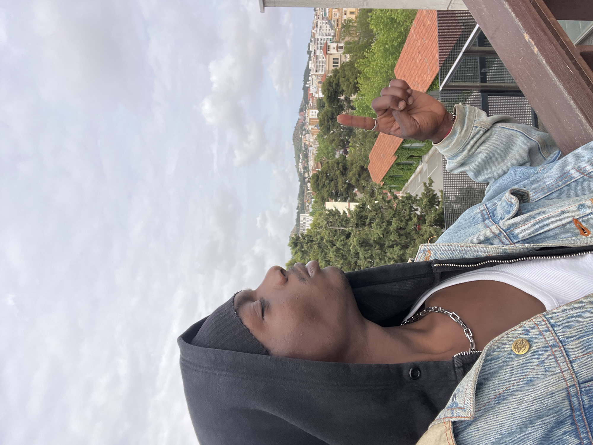 A low-angle medium shot depicts a person with brown skin, wearing a black hoodie, a white tank top, a silver chain necklace, and a denim jacket, standing outside. The person is facing the sky with eyes closed. The person is also extending a middle finger toward a city in the background. The background includes a cityscape, likely Barcelona, Spain, featuring various buildings, green trees, and other foliage, and a slightly cloudy sky. The foreground also contains parts of what appears to be a weathered wooden fence or railing.