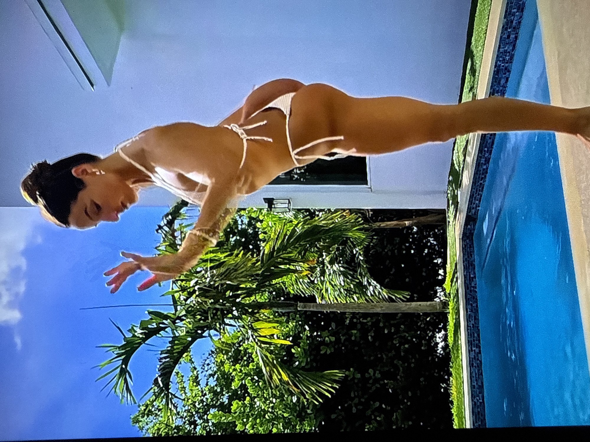 The scene is situated in Varna, Bulgaria, and depicts an outdoor setting under a clear, bright blue sky with sparse white clouds. A fair-skinned adult female, appearing to be in her 20s or 30s, stands in profile facing left. Her dark hair is pulled into a neat bun on top of her head. She wears a white string bikini, featuring a thong-style bottom, and is barefoot. Her hands are raised to chest height, palms forward with fingers slightly spread, and her fingernails are painted red. Gold hoop earrings adorn her ears, and multiple bracelets are visible on her right wrist. Her body is slightly bent forward at the waist, and her gaze is directed downwards or slightly ahead. She stands on a light-colored paved surface at the edge of a rectangular swimming pool filled with clear, blue water. The pool features a dark blue mosaic tile border and a light-colored coping. Behind the pool, a strip of vibrant green grass precedes a dense array of tropical vegetation, including several tall palm trees with slender trunks and abundant green fronds, and various dark green bushes. To the right of the frame, a portion of a modern, minimalist white building with a flat roofline and a visible overhang is present. A dark wall sconce is mounted on the building's exterior to the left.