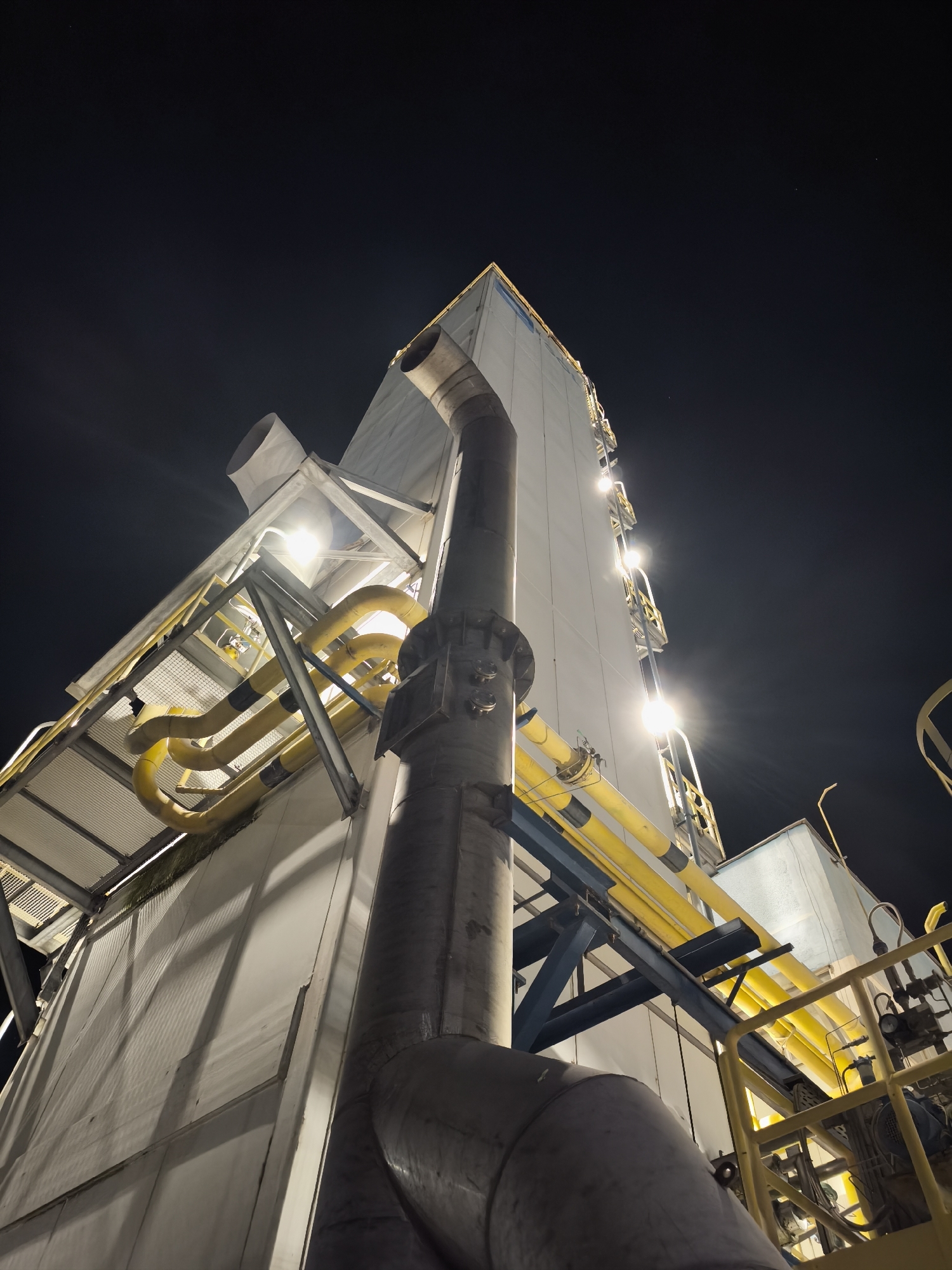 An industrial structure is depicted from a low-angle, worm's-eye perspective at night. The structure is predominantly white with visible vertical paneling, and it is illuminated by bright overhead lights, creating strong contrasts and shadows. Several large, yellow pipes, possibly conduits for gas or liquids, snake across the exterior of the building, supported by blue metal beams and a yellow railing system. A large, gray pipe is prominently featured in the foreground, curving upwards and to the right. The sky above is a deep, dark blue, with a few faint stars visible. The scene suggests an active industrial or processing facility.