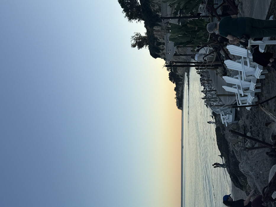 The image depicts a coastal scene in Malibu, United States of America, during dusk or late afternoon. The sky above transitions from light blue to a warm orange-yellow near the horizon, reflecting softly on the calm ocean surface. A rocky cliff, topped with green vegetation and trees, anchors the right side of the frame, overlooking the water.
In the left foreground, a woman in a dark shirt and blue cap is partially obscured while bending towards the rocky ground. Further along the shoreline, two silhouetted individuals and a lighter-colored figure are visible near the water's edge. A small, dark boat is positioned on the distant ocean.
In the midground, a row of five white Adirondack chairs are arranged on a sandy-rocky area, facing the ocean. Adjacent to these chairs, a wooden boardwalk with rope railings extends along the base of the cliff towards an outdoor dining or seating establishment. This establishment, situated at the base of the cliff, features suspended string lights and large leafy plants. Patrons are visible seated at tables and chairs within this area. In the immediate right foreground, an individual with grey hair, wearing a dark green short-sleeved top, stands observing the scene, with another person seated directly in front of them.