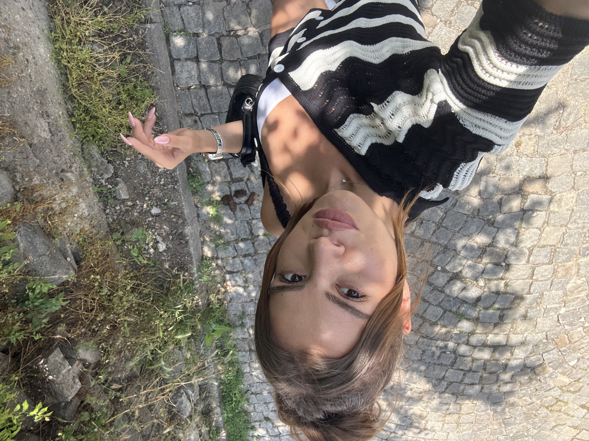 The image is a high-angle selfie of a young woman standing outdoors on a sunny day. She has medium-brown hair pulled back, with some strands framing her face, and a neutral expression as she looks directly into the camera.
She is wearing a black and white knitted or crocheted cardigan with a wavy, horizontal stripe pattern, worn open over a white top with a thin black strap visible on her left shoulder. On her left wrist, she wears a silver-toned watch and a decorative bracelet with multiple rows of small, clear stones. A delicate silver necklace with a small circular pendant is visible around her neck. Her nails are long, pointed, and painted a light pink color. A black handbag with silver hardware hangs on her left side.
The setting consists of gray cobblestones underfoot, bordered on the left by a concrete curb and an area of dirt and wild green and brown vegetation. The bright sunlight casts prominent shadows on the cobblestone ground. There is no visible text in the image.