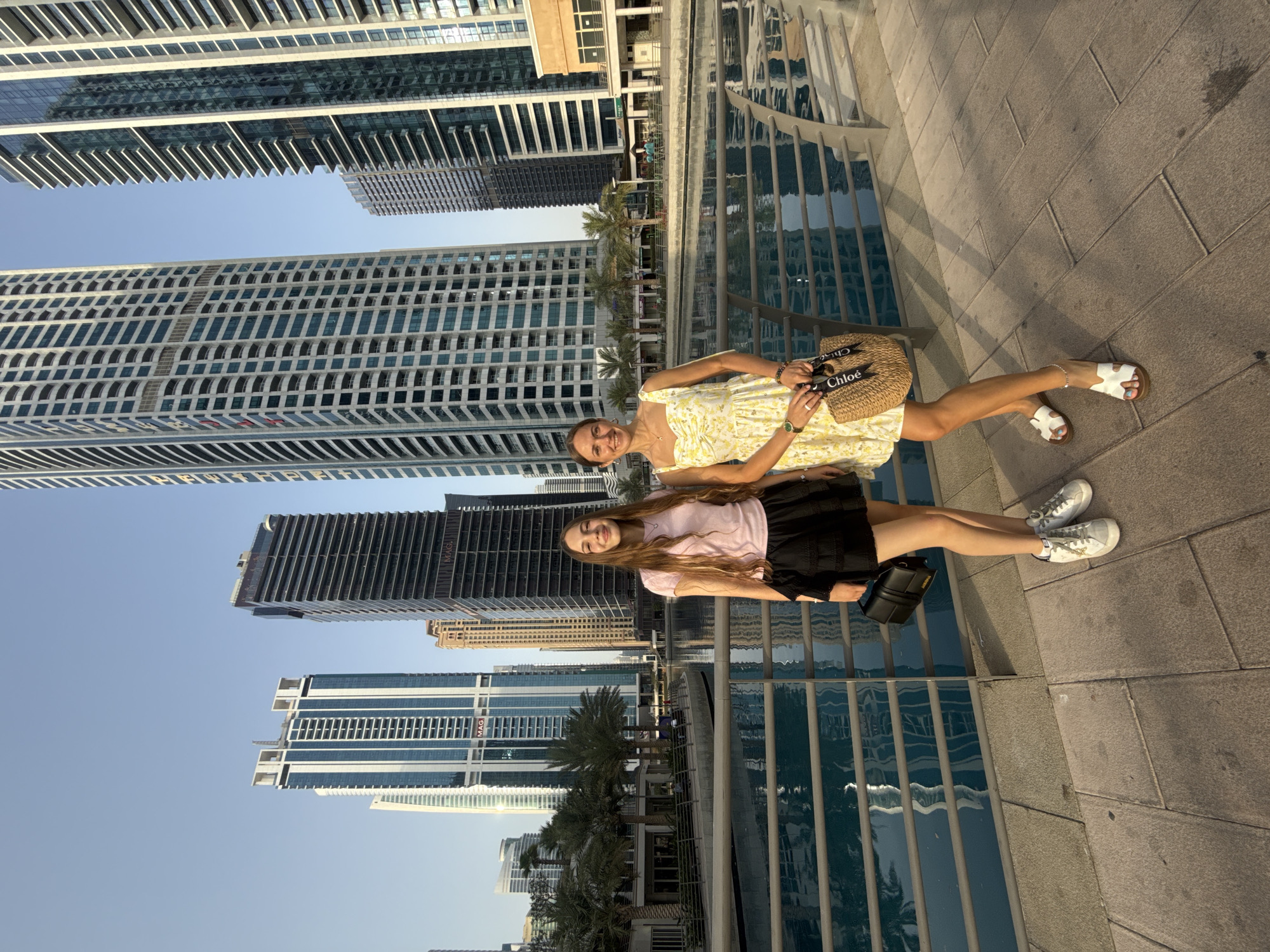 Two women stand on a paved walkway in front of a body of water with a railing between them and the water. The woman on the left has long brown hair and wears a pink t-shirt and a black skirt. She holds a black purse in her left hand. The woman on the right has dark hair and wears a yellow floral dress. She holds a woven bag in her right hand. Both women are smiling and looking at the camera. Behind them, several modern skyscrapers tower over palm trees and low-rise buildings. The water reflects the buildings and the sky. The scene appears to be in Dubai, United Arab Emirates, possibly near the Dubai Water Canal given the architecture and water features. The lighting suggests it is late afternoon or early evening.