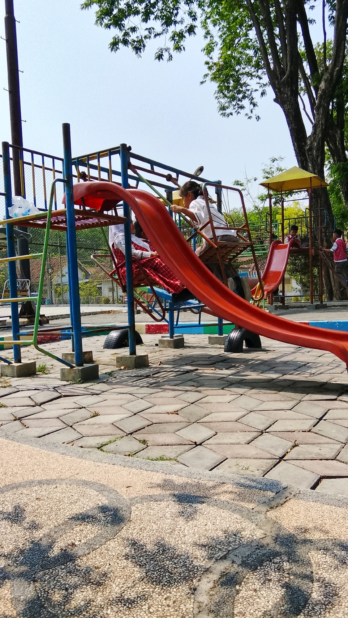 A bright, sunny day is depicted at a playground in Tlogobendung, Indonesia. The playground features a red slide that curves downwards from a metal structure with blue and yellow supports. Two children, wearing white shirts and red and white checkered skirts or shorts, are visible on the playground. One child is seated on the lower part of the slide, while another is positioned further up, appearing to be interacting with the slide or the equipment. Other children can be seen in the background, some climbing on the structure, and one appears to be on a smaller red slide. The ground of the playground is paved with interlocking gray and beige stones, with some patches of grass peeking through. Large trees with green foliage provide shade in the background, and a residential house can be seen beyond the playground fence. The overall scene suggests a lively and active environment for children.