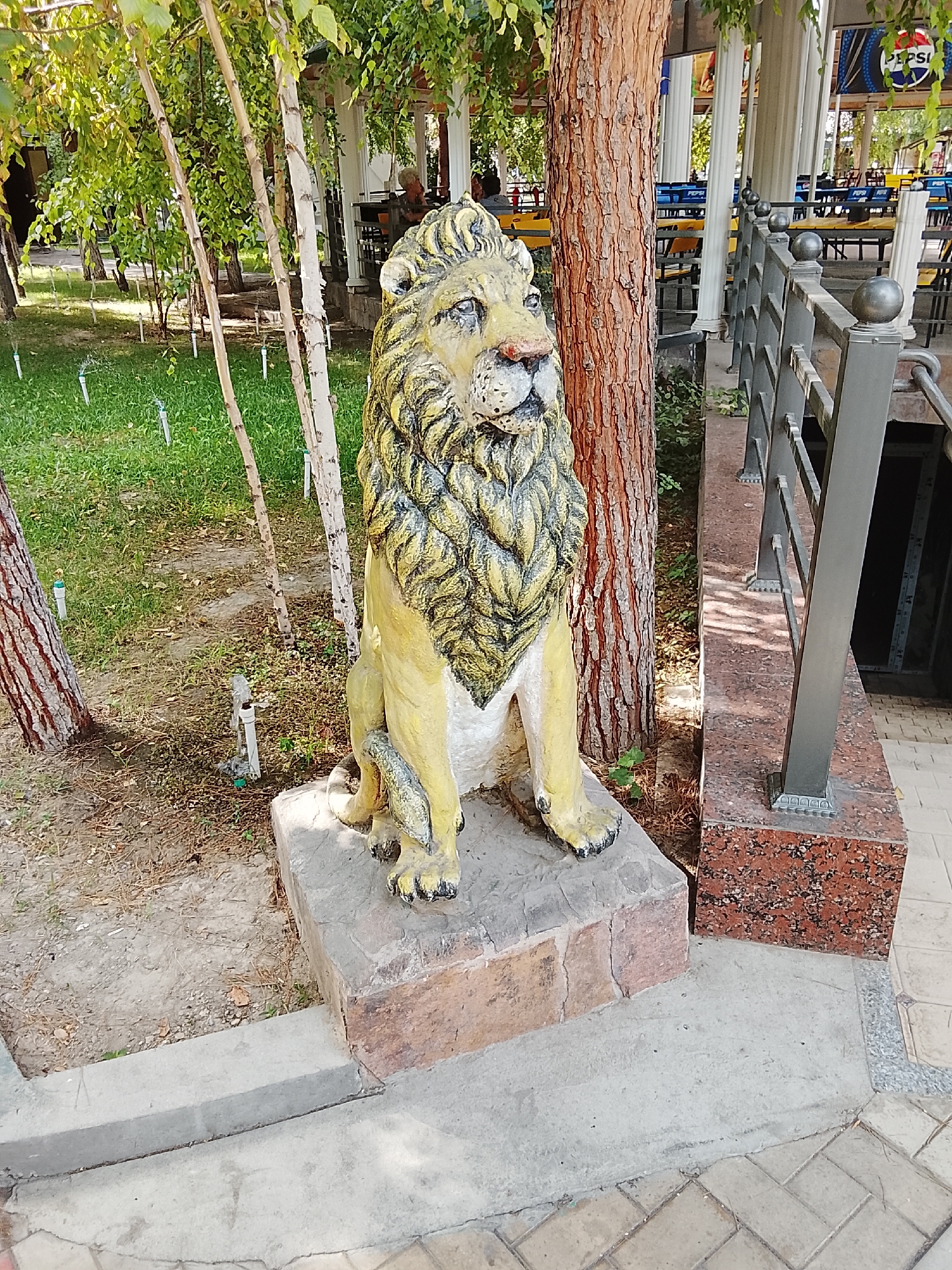 The image shows a concrete statue of a lion sitting on a stone pedestal in what appears to be an outdoor seating area of a cafe or restaurant. The statue is painted yellow with black accents, and its mane is depicted with textured lines. Behind the statue, there is a large tree trunk, and further in the background, tables and chairs are visible, some of which are covered by a canopy supported by white pillars. A railing with metal posts separates the seating area from a downward sloping path. A Pepsi advertisement banner is visible in the far background. The ground is a mix of concrete and paving stones, with some patches of grass and dirt. The overall setting suggests a public recreational space within Fergana City, Uzbekistan. There are no people clearly visible in the foreground.