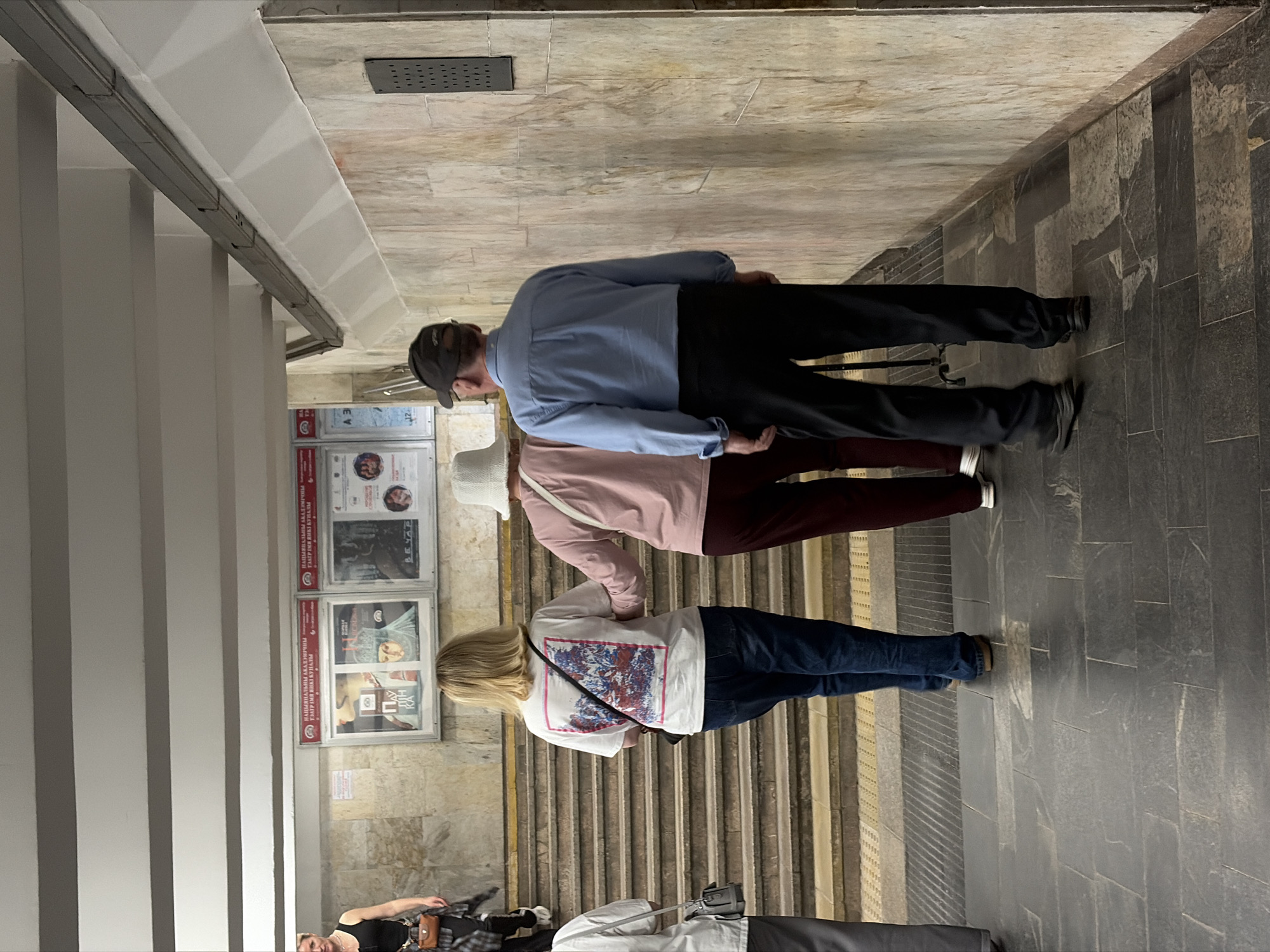The image captures a scene within what appears to be a subway station. A group of three people are walking down stairs. The first person has on a blue long sleeve shirt, black pants, and a black baseball cap. The second person has a light pink long sleeve shirt, maroon colored pants, and a white bucket hat. The third person has a white t-shirt, blue jeans, and blonde hair. A set of framed posters hang on the wall to the left of the stairs. A person who looks to be homeless is at the bottom of the stairs. The station walls are tiled in beige, while the stairs are a mix of beige and gray stone. The scene has an urban feel, with the architecture and the presence of people suggesting a busy public space. The city is unknown.