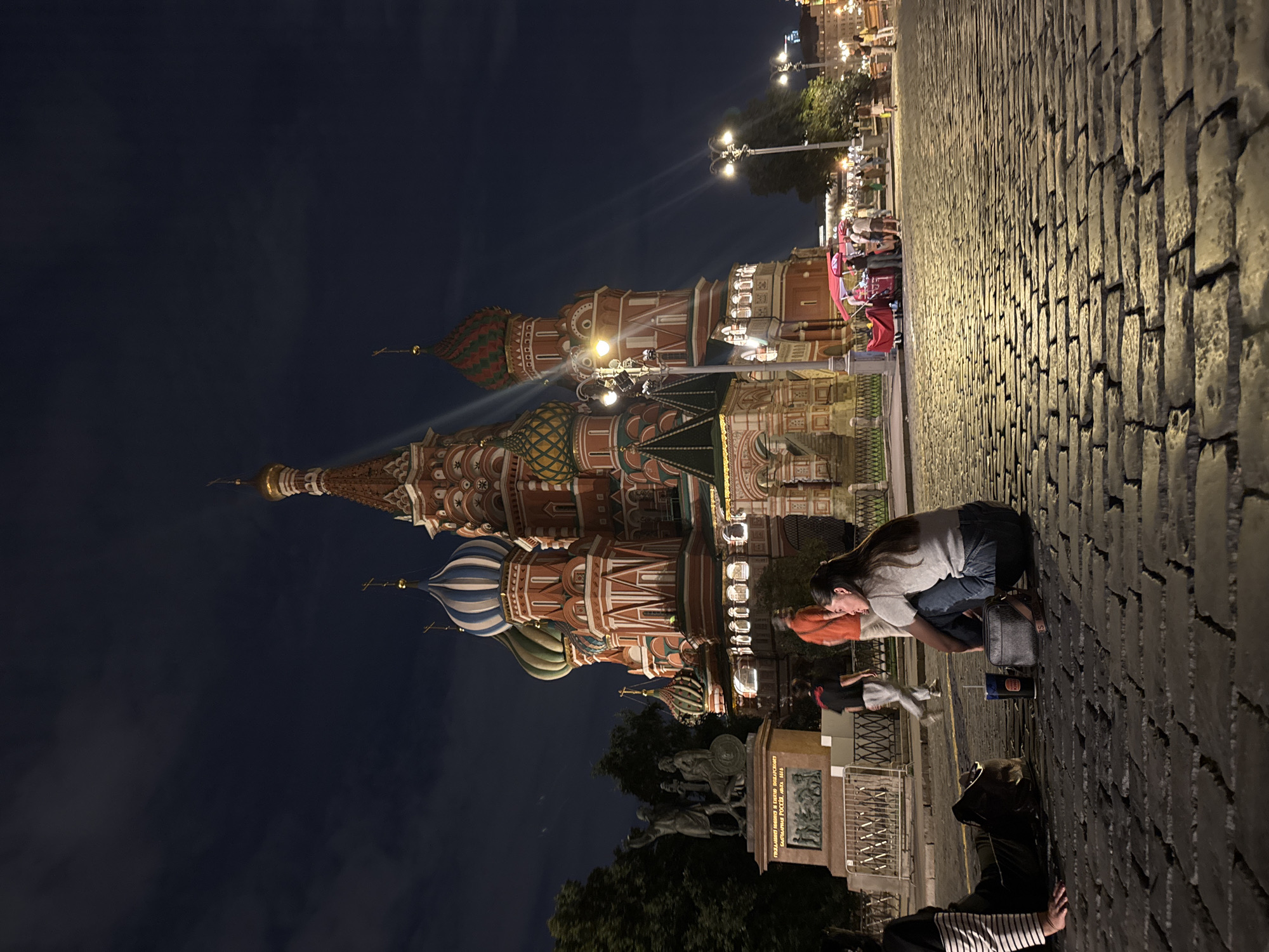 The image captures a nighttime view of Saint Basil's Cathedral in Moscow, Russia, located on Red Square. The iconic onion domes and intricate architecture of the cathedral are illuminated against a dark, cloudy sky. In the foreground, a cobblestone plaza is visible, with a low-angle perspective emphasizing the texture of the stones.
Several people are present in the scene. In the mid-ground, a couple is sitting on the cobblestones, facing away from the camera and towards the cathedral. The woman is wearing a grey t-shirt and blue jeans, while the man is wearing a light-colored t-shirt and dark pants. They appear to be resting or observing the surroundings.
To the left of the couple, a person is lying down on the cobblestones, with their legs crossed and their upper body partially visible. This person is wearing a striped long-sleeved shirt. A blue and black beverage cup and a small grey bag are placed on the ground near the seated couple.
In the background, other individuals are walking and gathered near a stall or kiosk with red and pink coverings, suggesting a vendor or food stand. Statues and trees are also visible to the left of the cathedral, and streetlights cast a warm glow on the plaza, illuminating the scene and creating visible light rays. The overall atmosphere is one of a vibrant public space at night.