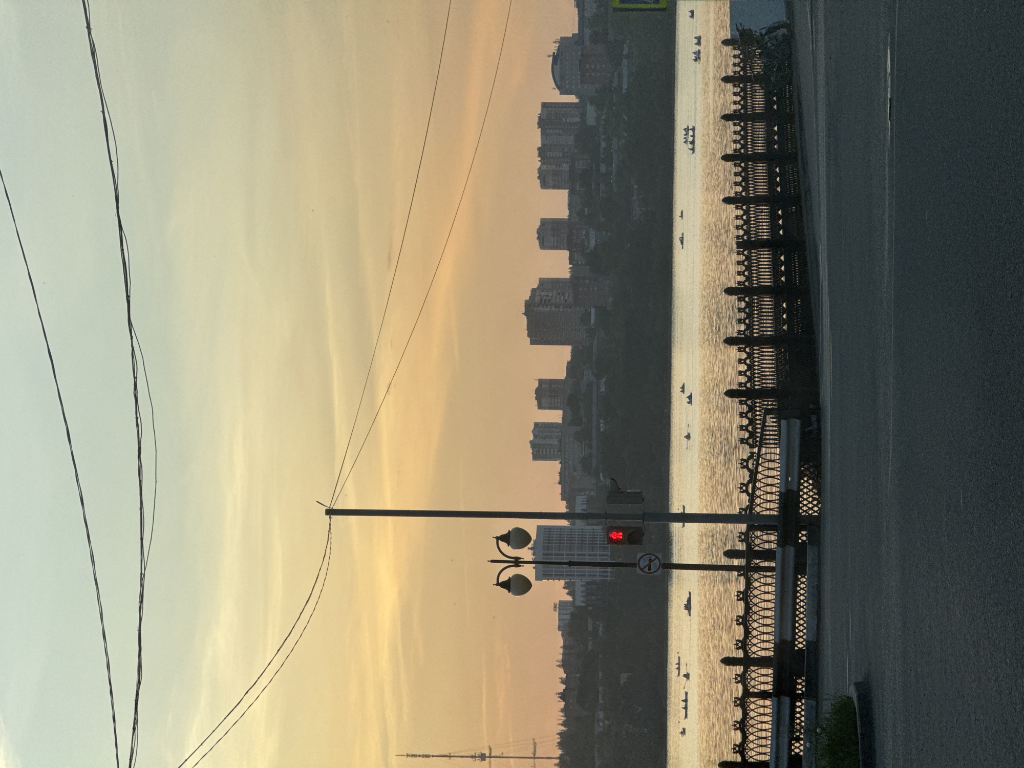 The image shows an evening cityscape of Izhevsk, Russia, with a river separating the viewer from a row of buildings. The sky is a mix of yellow and pale blue, likely due to the sunset. Power lines run vertically on the left side of the image. A lamppost and a traffic light with a red light are positioned near a metal fence and a road on the right. Several boats can be seen on the river.