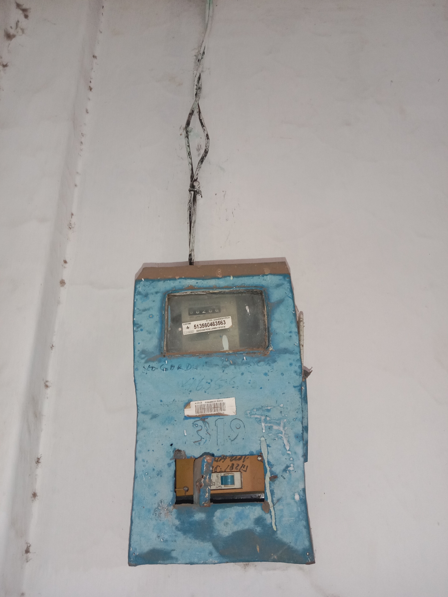 The image shows a close-up, eye-level view of a blue electrical meter box mounted on a white, textured wall. The meter box is weathered, with peeling paint and some grime. A glass window on the meter box reveals a digital display with the number "00200". Below the window, a label reads "513560463563". Further down the box, the number "319" is visible, along with a small brown panel with a yellow switch and a date stamp that appears to read "18/2/19". Electrical wires are visible entering the top of the meter box. The overall impression is of an older, possibly rural, installation. The location is Pohsangit Leres, Indonesia.