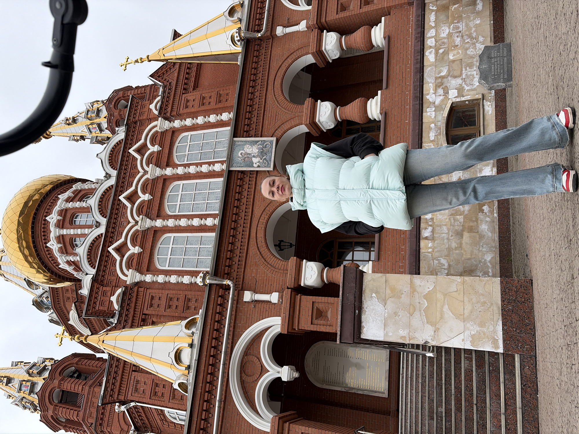 A single woman stands centered in the foreground, facing the camera directly, with her hands in the pockets of her light blue sleeveless puffer vest. She is wearing a black long-sleeved shirt, light-wash flared blue jeans, and red and white sneakers.
Behind her stands St. Michael's Cathedral (Mikhailovsky Cathedral) in Izhevsk, Russia, a large red brick Orthodox church with multiple golden onion domes and spires. The cathedral's facade features intricate white decorative trim, arched windows, and a prominent icon depicting Saint George slaying the dragon positioned above a central entrance. To the left of the woman, a set of granite-like stairs with a metal handrail leads up to the cathedral's entrance, adjacent to which a large plaque with Russian text is affixed to the wall. The lower section of the cathedral's facade displays a mix of brickwork and light-colored stone with visible staining and wear.
The ground is paved with reddish-brown cobblestones. A rough-hewn stone with Cyrillic text is partially visible on the ground to the right of the woman. The sky is overcast. A black, curved object is visible at the top right of the frame, partially obstructing a view of one of the cathedral's spires.