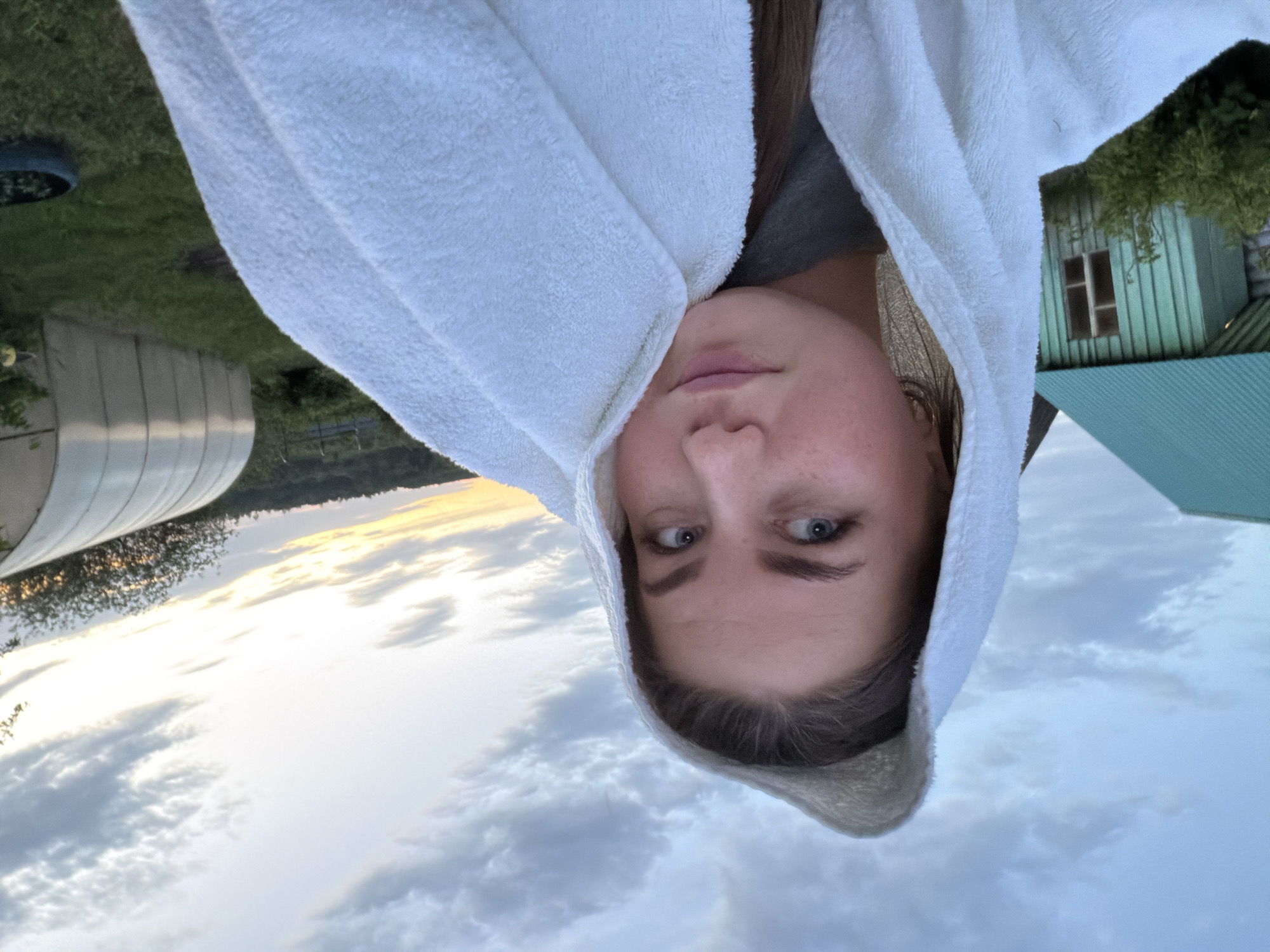 A young person with fair skin and blue eyes is looking directly at the camera, wearing a white hooded towel. The background shows a rural setting at sunset. There is a greenhouse to the left, a small green building with a window to the right, and a cloudy sky. The location is Уральское, Russia.