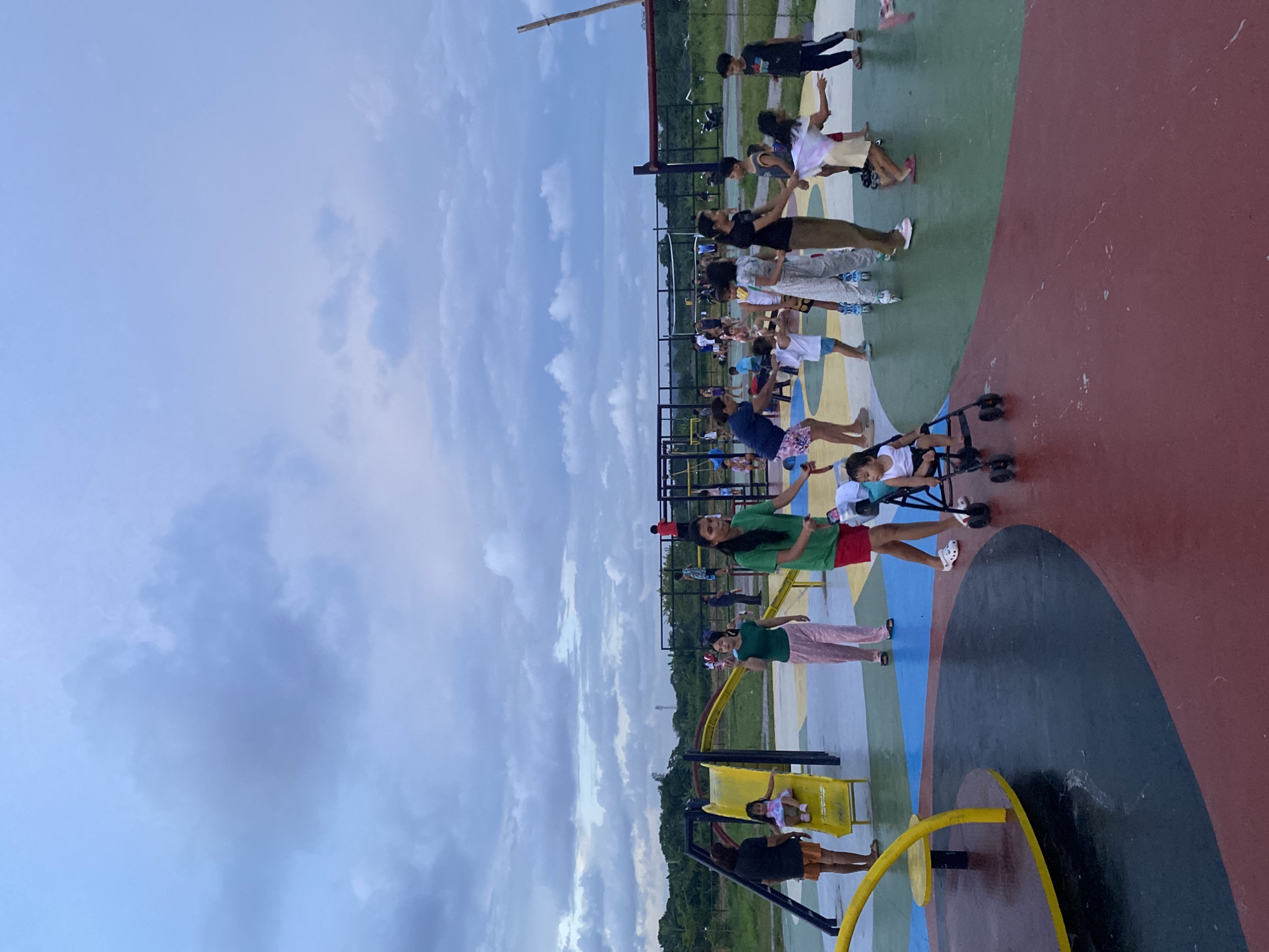 The image depicts an outdoor playground scene in Davao City, Philippines, characterized by a vibrant, multi-colored rubberized surface with distinct zones of red, green, blue, and yellow. In the foreground, a yellow, circular merry-go-round structure is partially visible on the left. Several people, including adults and children, are engaged in various activities. On the left, a young girl in a pink dress is on a yellow slide. A woman in a black shirt and orange shorts stands nearby. To the right of the slide, a woman in a green shirt and pink pants walks towards the viewer. In the center, a toddler sits in a walker-like stroller. Further back, a group of children and adults are scattered across the playground. Some children are playing near a structure with yellow bars and what appears to be a volleyball net in the background. The sky above is overcast with dramatic, dark gray clouds. The environment suggests a public recreational area, possibly within a park or open space.