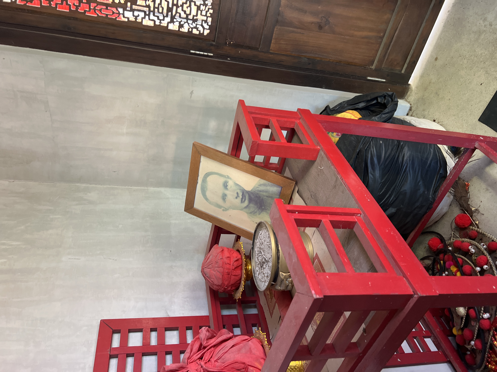 The scene depicts the interior of what appears to be a storage area or neglected corner of a building. The location is identified as Baan Hua Thanon, Thailand. The dominant feature is a red, tiered structure, possibly a dismantled altar or storage unit. Various items are placed haphazardly on and around it, including a framed portrait of a Caucasian individual, an incense burner, and fabric-covered objects, some red and some gold. There is a black trash bag near the structure. Other items include assorted decorative objects with red pom-poms and white beads. The walls are painted in a neutral tone. A section of an ornate wooden door or screen is visible at the top of the frame. The items give the impression of an area where sacred or culturally significant objects have been stored or discarded.
