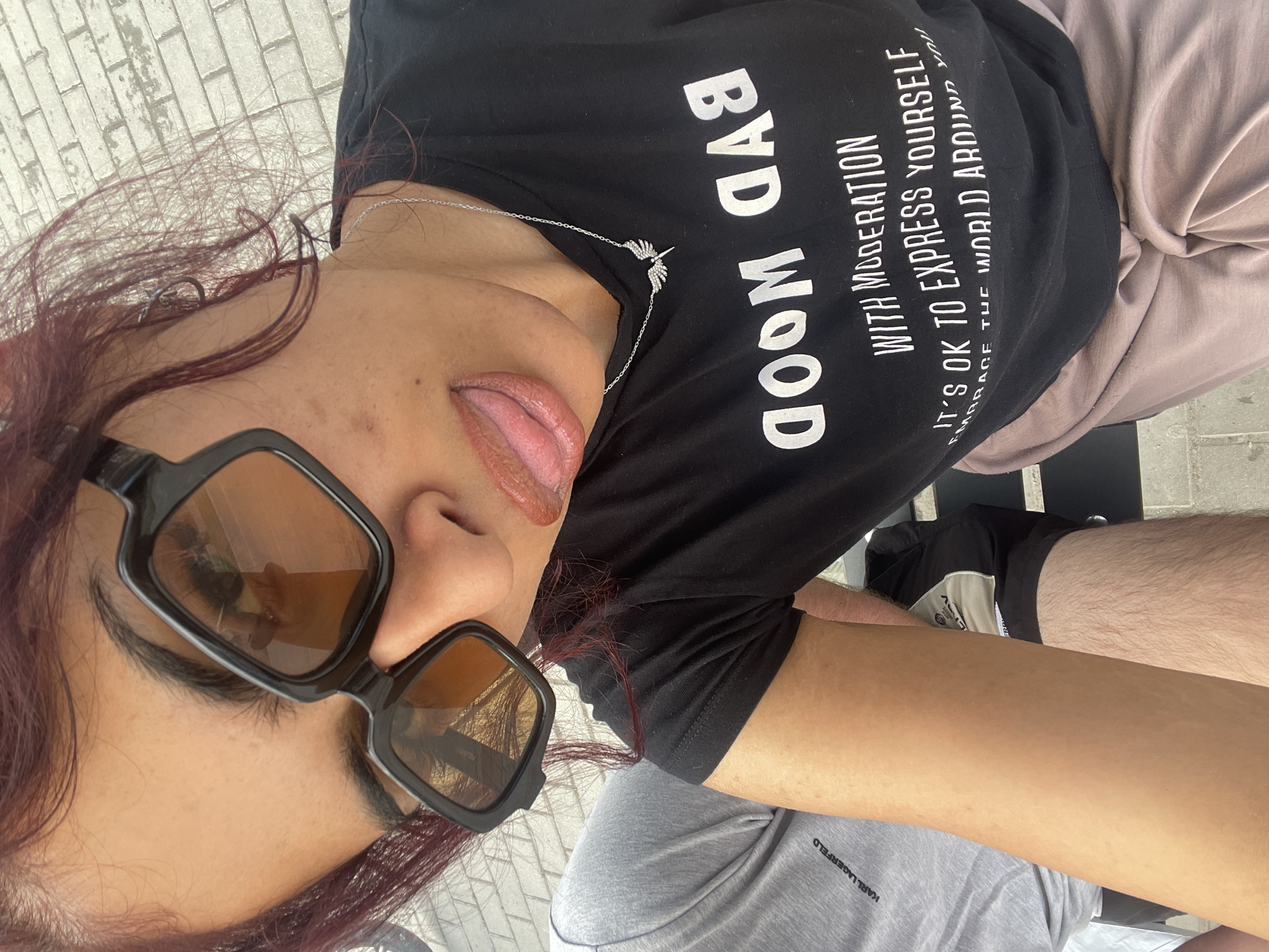 An overhead selfie features a person with red hair, wearing black square-framed sunglasses with brown lenses, sticking their tongue out. A silver chain necklace with a small winged pendant rests on a black t-shirt. The t-shirt displays reversed white text, partially visible, which reads: "BAD MOOD WITH MODERATION IT'S OK TO EXPRESS YOURSELF EMBRACE THE WORLD AROUND YOU." The person wears light brown or taupe pants. To the left, a leg from another individual is visible, clad in light grey shorts or pants with a "KARL LAGERFELD" logo near the hem. The ground is paved with light-colored, rectangular bricks or pavers. The scene is outdoors in Unknown, Unknown.