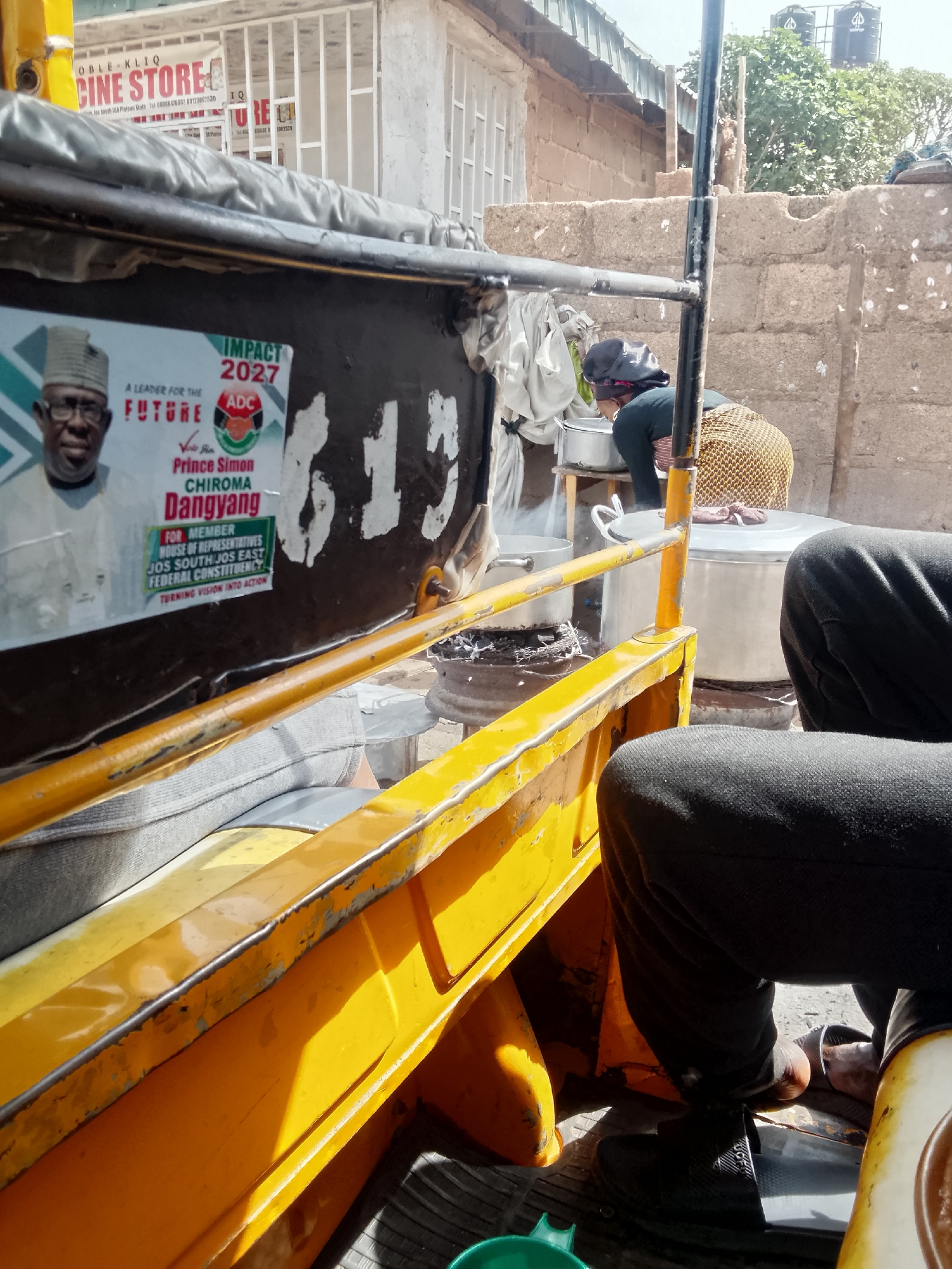 Captured from inside a "keke napep" (auto-rickshaw) in Bukuru, Nigeria, the image presents a scene of daily life. In the foreground, a section of the yellow keke napep frame is visible, along with the legs of a seated passenger wearing black trousers and sandals. The back of the keke displays a political poster with the inscription "IMPACT 2027," featuring a photograph of Prince Simon Chiroma Dangyang, a candidate for the House of Representatives. Through the keke's open side, a woman is observed cooking outdoors, tending to large pots set over an open fire. The backdrop consists of a modest, unfinished building constructed with concrete blocks, as well as a store. Overhead, water tanks are visible on the rooftops, indicating urban infrastructure.
