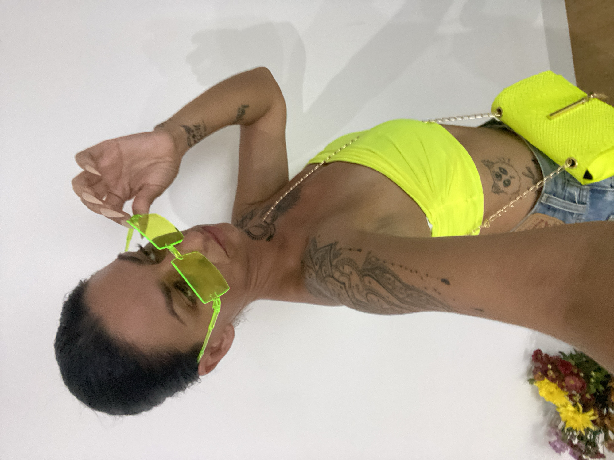 A woman is posing for a selfie in a room with a white wall. She has dark hair pulled back and is wearing neon yellow sunglasses, a neon yellow tube top, and denim shorts. A matching neon yellow handbag with a gold chain strap is slung over her shoulder. She has tattoos on her arms and torso, including one of a cat with multiple eyes on her stomach. To the left of the frame, a bouquet of flowers in purple, yellow, and red hues is visible. The image does not contain any information about the city of Mũi Né, Vietnam.