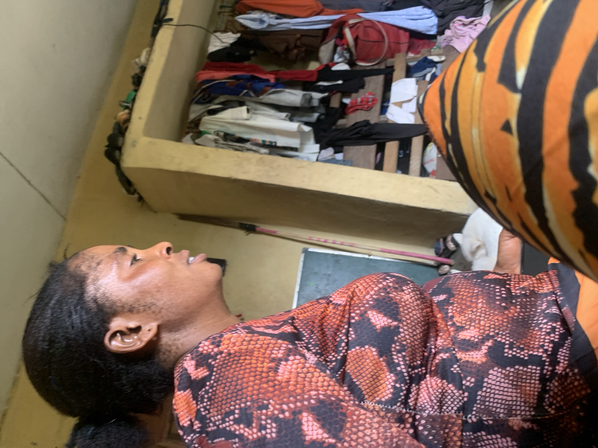 The image shows a person lying down indoors with their head tilted back. They are wearing a snakeskin-patterned shirt and a striped orange and black garment. The environment appears to be a cluttered room with clothes piled up in a corner. The location in the city of Aba, Nigeria is uncertain; there are no objects or elements in the image that can be used to discern a specific location.