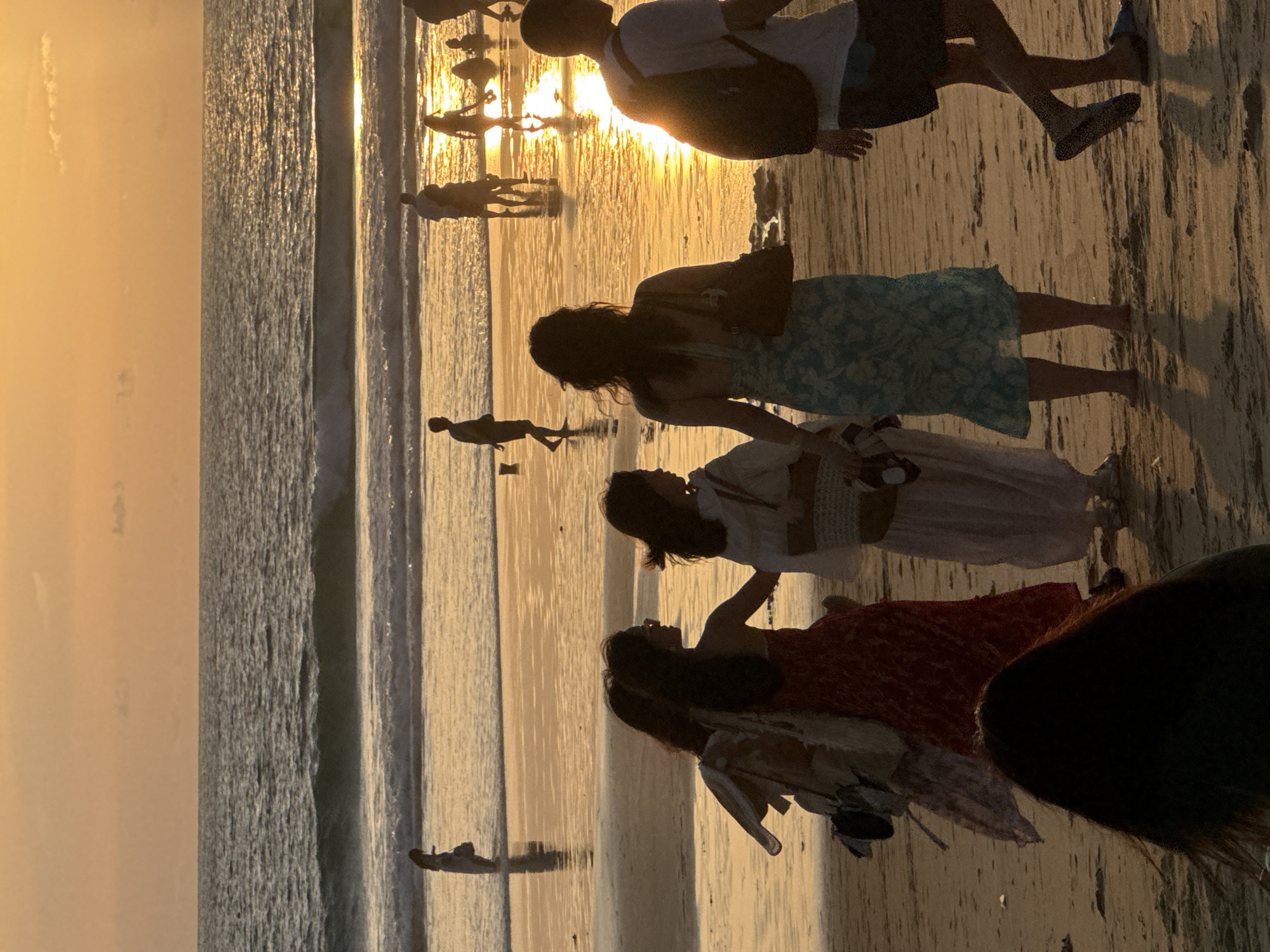 At sunset on Karon Beach in Karon, Thailand, a group of people are walking along the wet sand near the ocean. The sky is a warm orange and yellow, reflecting on the water and the wet sand. In the foreground, several individuals are walking away from the camera, their silhouettes dark against the bright backdrop. One person in the foreground is wearing a light-colored patterned dress and carrying a dark bag. To their left, another person is wearing a red patterned dress. Further back, a group of people are visible further out on the beach. The ocean has gentle waves, and the sun is low on the horizon, creating a bright reflection on the water's surface. The scene conveys a peaceful evening atmosphere on a tropical beach.