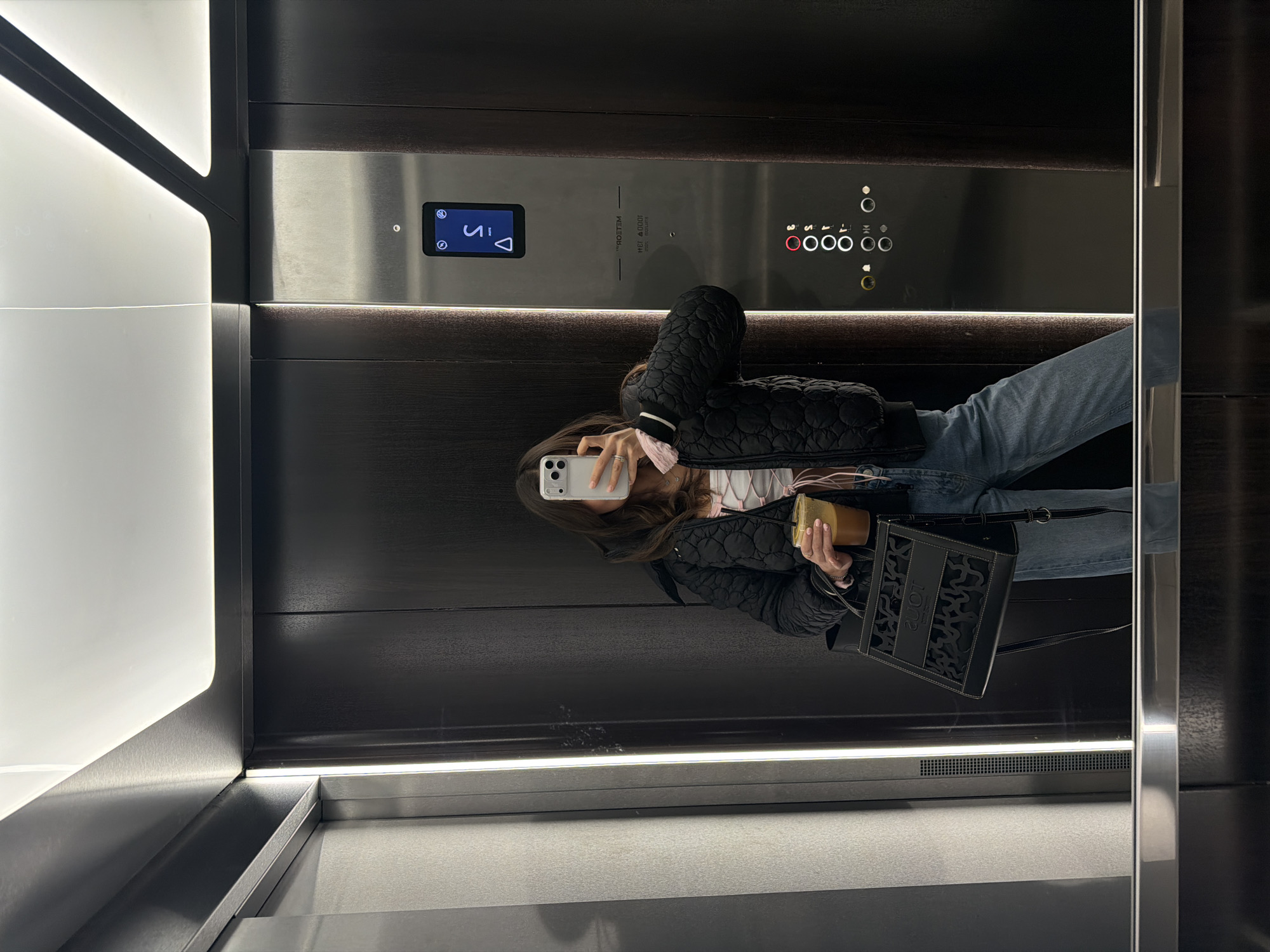 A person is captured in a mirror selfie inside a modern elevator, facing the reflective door. The subject, likely female, wears a black quilted jacket over a light pink top and blue jeans. Her face is obscured by a white smartphone (an iPhone with three cameras) which she holds up to take the photo. In her left hand, she holds a transparent plastic cup with an iced beverage. A black tote bag with a distinctive cut-out design, partially revealing "LOUIS" lettering, hangs from her shoulder.
The elevator interior features sleek, dark wood-grain panels contrasted with bright, linear LED strip lighting along the walls and floor, creating a contemporary ambiance. The main control panel, situated on a brushed metal surface, includes a digital display showing "S" and an upward arrow, indicating the elevator's current status or direction. Below this, various circular buttons are visible: "3", "2", "1", "0", "M", "B", along with symbols for door control, a bell, and an emergency stop. Mirrored text on the panel, reversed in the reflection, reads "WELCOME" and "1000 кВ" (likely indicating a 1000 kg capacity) and "МЕRТЕLМ" (possibly a brand name, reversed). The scene depicts an everyday moment within an urban setting in Krasnoyarsk, Russia.