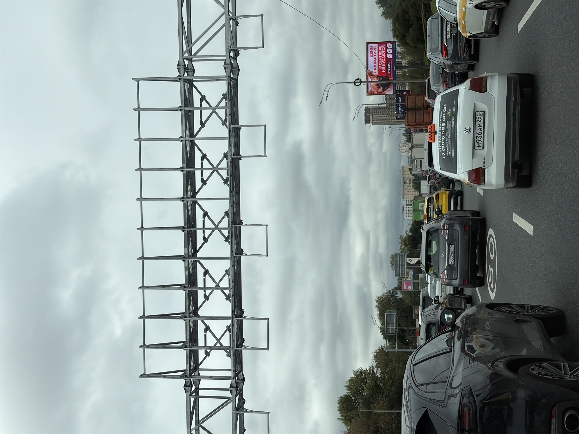 The image depicts a traffic jam on a cloudy day in Moscow, Russia. The foreground shows the rear of a black SUV, followed by several other cars including a white Volkswagen Polo taxi with Russian license plate M936AM 250. In the background, buildings and trees are visible. A large billboard advertising apartment prices is also present. The scene appears to be a typical urban traffic situation.