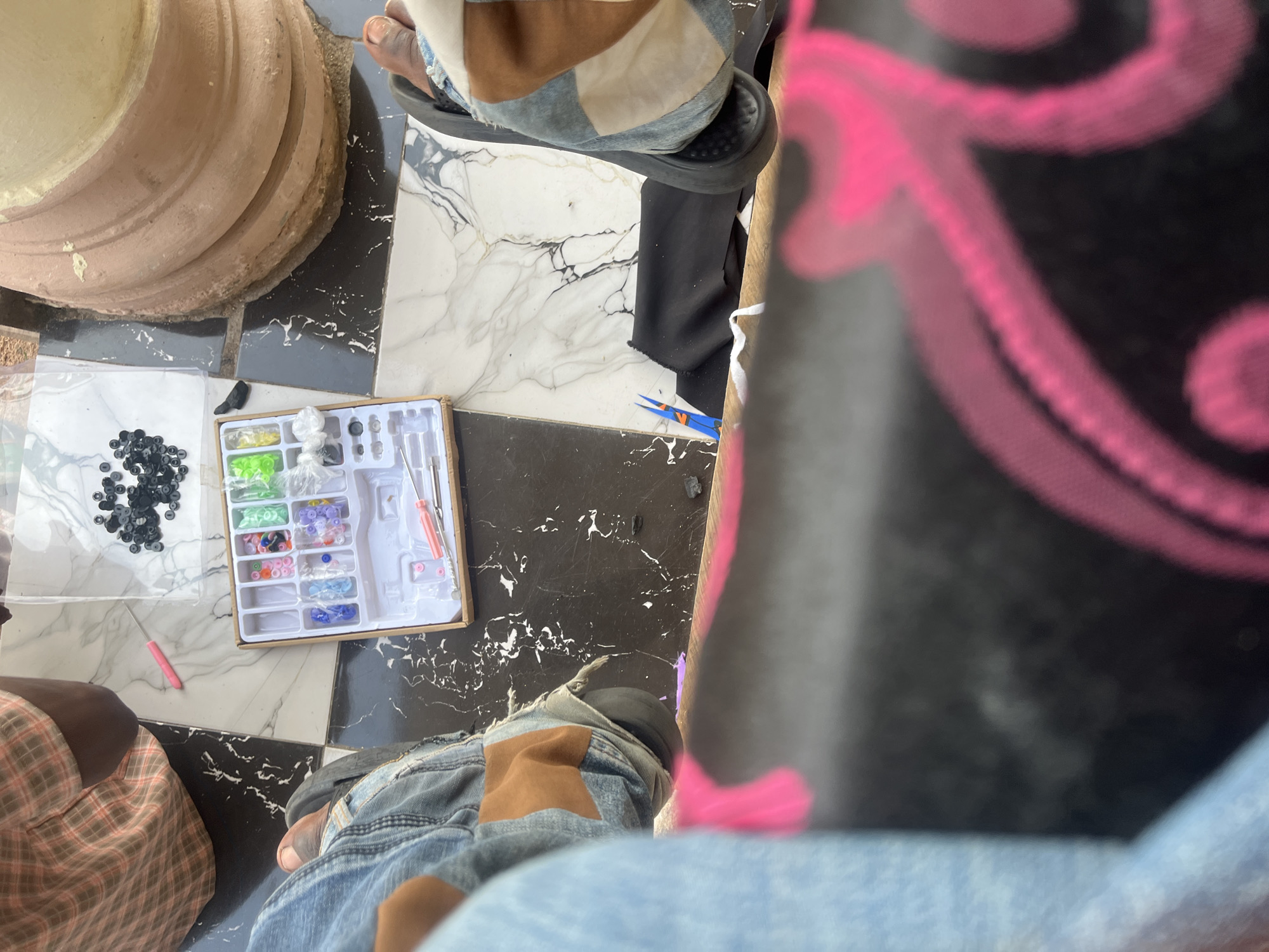This media file shows a top-down view of a person sitting on a marble-patterned floor, engaged in a craft activity. The subjects include the person's legs, clad in jeans with a suede patch on one knee, and their feet wearing sandals. The scene is laid out with various craft supplies, including a kit of colorful beads in a white organizer, a bag of black beads, and a pink crafting tool. A large, round, terracotta-colored object, possibly a pot or planter, is visible in the upper left corner. The background features a black and white marble-tiled floor. The lighting suggests it is daytime. There are no visible text elements in the image. The overall impression is one of a casual, hands-on activity taking place in a residential or outdoor setting in Jalingo, Nigeria.