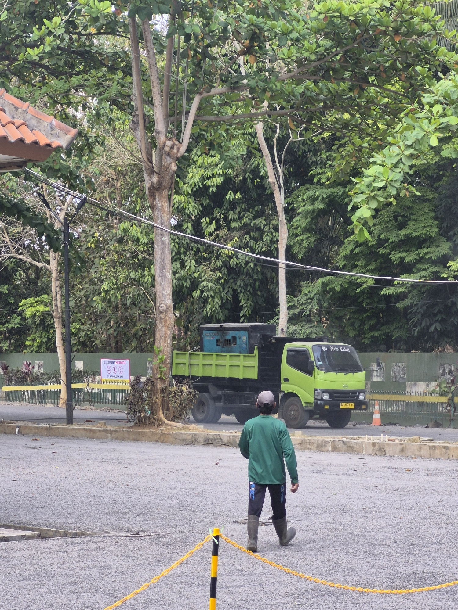 In Purworejo, Indonesia, a man wearing a green long-sleeved shirt, dark pants with blue accents, and grey rubber boots walks away from the viewer across a gravelly, open area. He has a light-colored cap on his head. In the background, a bright green dump truck with a black bed is parked. The truck has "Pt. New Calii" written on its windshield. The scene is set amidst lush green trees and foliage, with a partially visible red-tiled roof of a building to the left. Electrical wires are strung across the trees. A yellow chain barrier with black and yellow striped posts is visible in the foreground and extends into the distance. A sign with Indonesian text and symbols, including a red circle with a line through it, is posted behind a fence to the left of the truck.