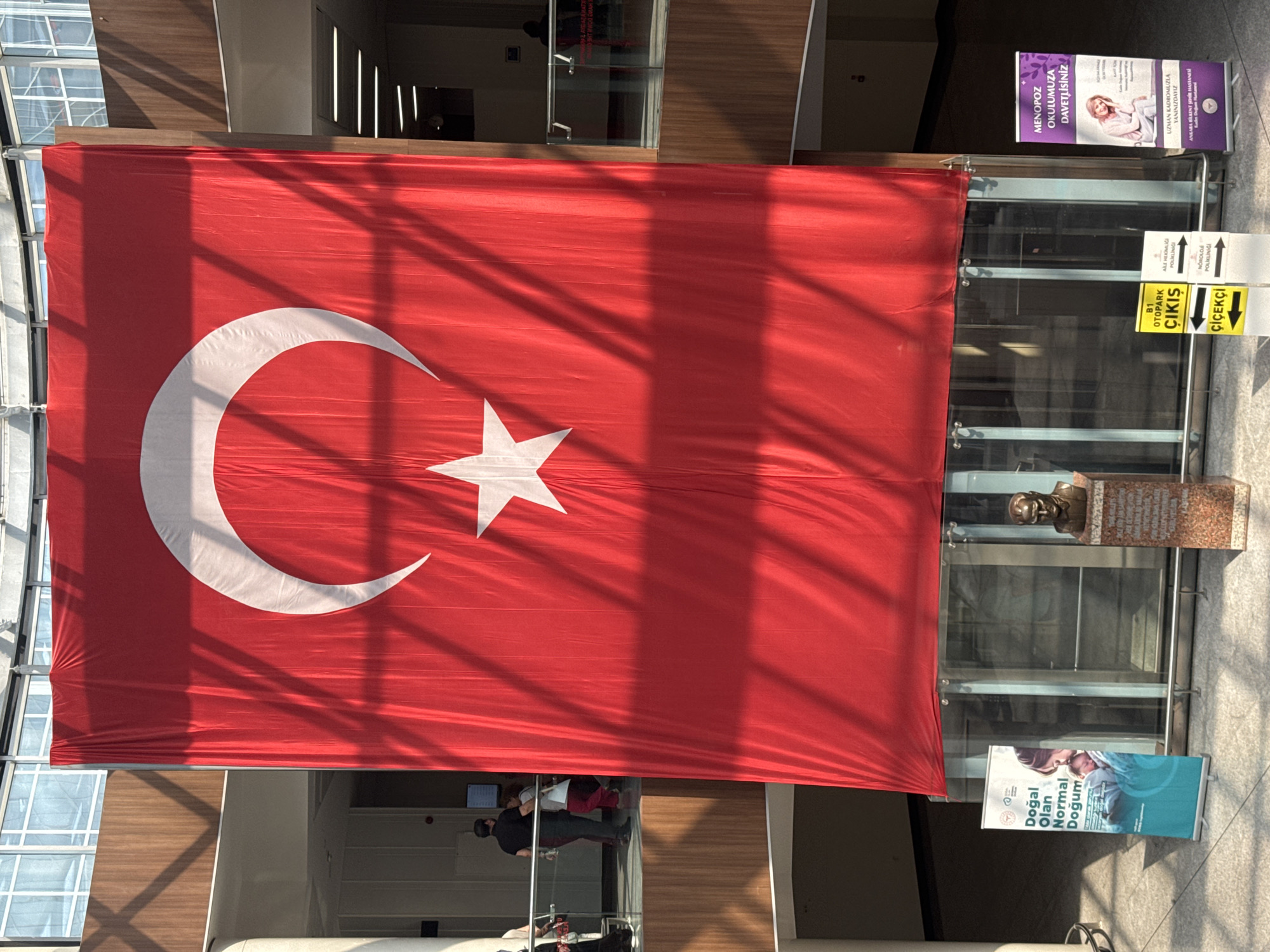 A large Turkish flag is displayed prominently, hanging from a ceiling and reaching down towards the ground. The flag features a white crescent moon and a white star on a red background. Below the flag, a bust of Mustafa Kemal Atatürk is mounted on a pedestal. To the left of the bust, a vertical banner with the text "DOĞAL OLAN NORMAL DOĞUM" (Natural Birth is Normal Birth) is displayed. To the right of the bust, another vertical banner reads "MENOPOZ OKULUMUZA DAVETLİSİNİZ" (You are invited to our Menopause School). Multiple directional signs are visible, including "B1 OTOPARK" (B1 Parking), "ÇIKIŞ" (Exit), and "ÇİÇEKÇİ" (Florist). In the background, people are seen walking on an upper level, and the environment appears to be an indoor public space with architectural elements such as wooden panels and large windows. The presence of the Turkish flag and the bust of Atatürk suggest a patriotic or memorial context, possibly within a public building in Çankaya, Turkey.