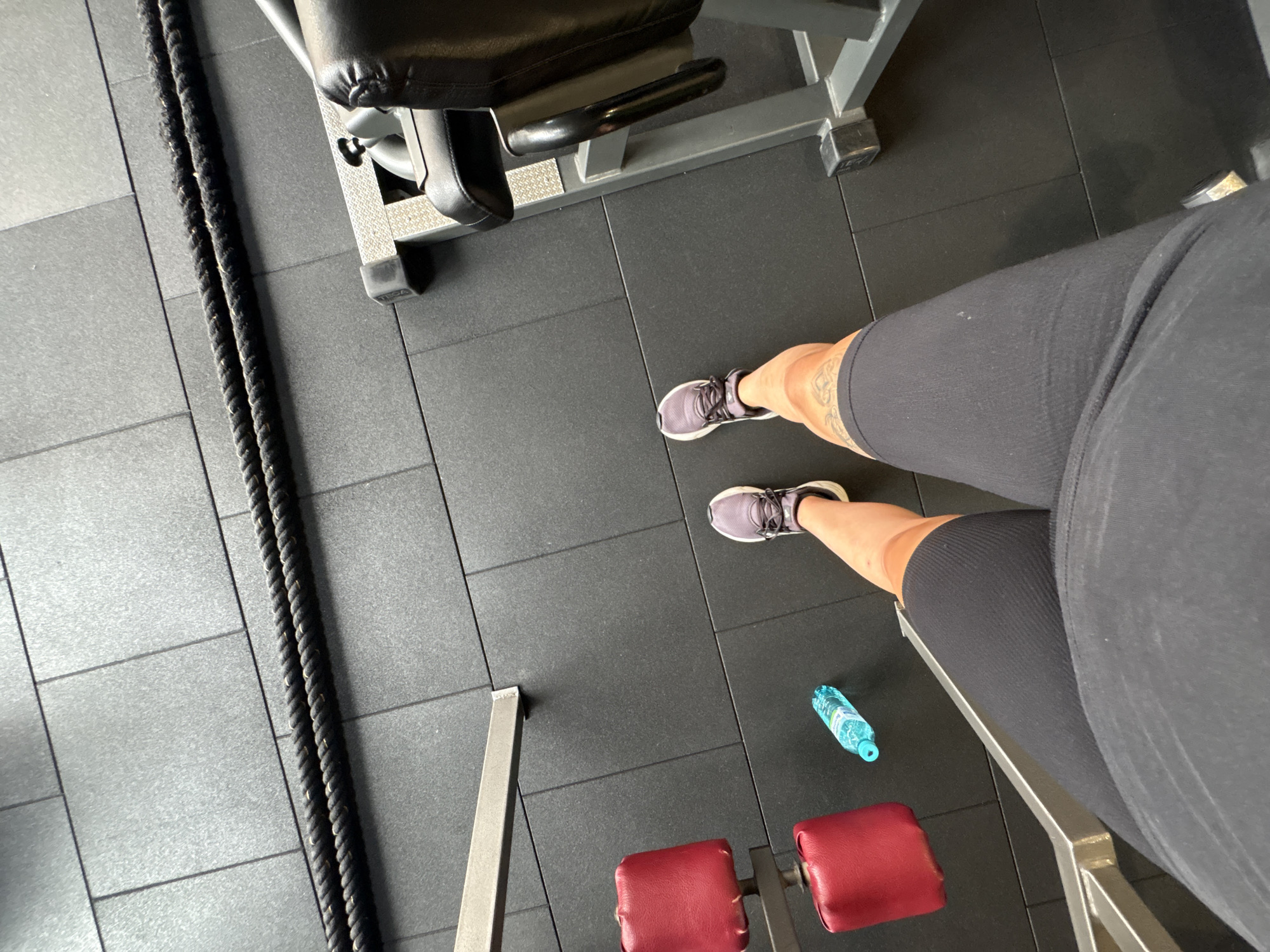 The image shows a person's legs and feet wearing athletic shoes and black shorts in a gym. Two red cushioned pads are visible on the left, attached to a metal structure. A blue water bottle lies on the black rubber gym floor. Two thick black ropes are positioned horizontally in the background. The gym floor is made of large, dark grey square tiles. The scene is observed from a high-angle perspective, looking down at the person's legs. The location is the city of Troyan, Bulgaria.