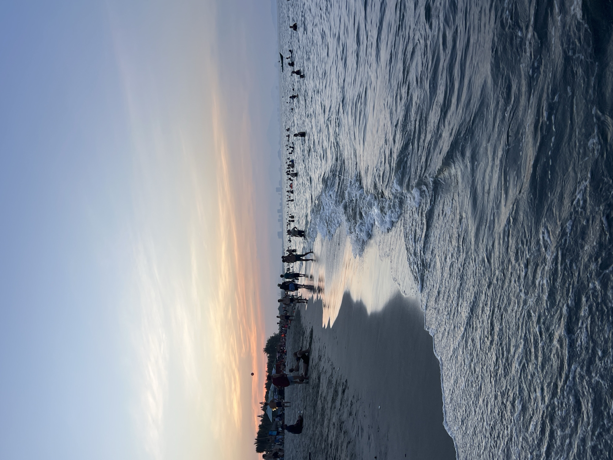 The image shows a beach scene in Hoi An, Vietnam at sunset. The sky is a gradient of pale blue and soft orange, with wispy clouds. The sea is calm with gentle waves lapping the shore. Numerous people are present on the beach and in the water, some walking along the sand, others swimming or wading. The beach is sandy with some vegetation and structures, possibly umbrellas or tents, visible in the background. In the distance, a boat is visible on the water, and a city skyline can be seen across the horizon. The overall atmosphere is one of leisure and recreation.