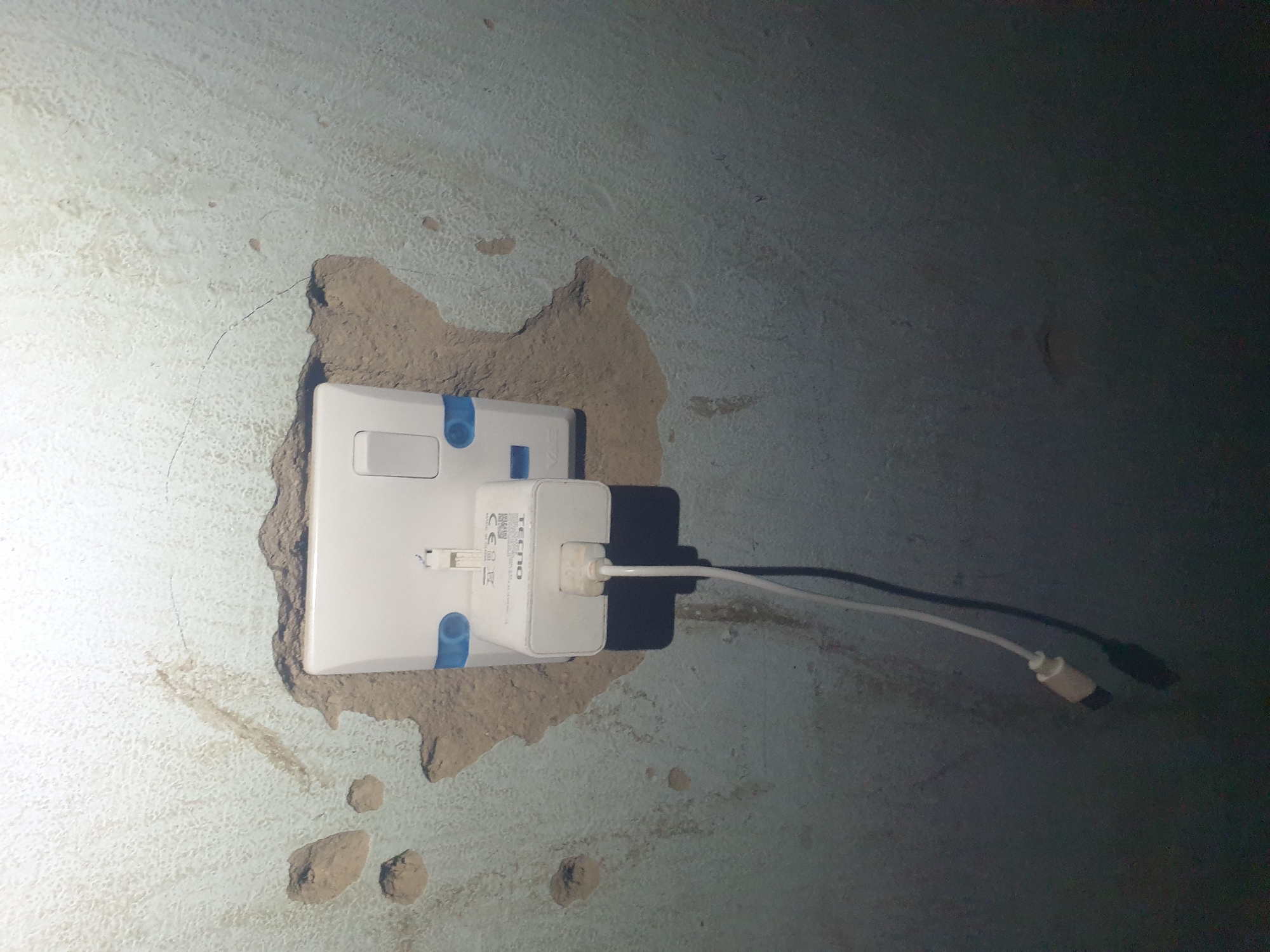 The image shows a close-up of a white electrical outlet on a textured, light-colored wall with some water damage and cracks. A white Tecno brand phone charger is plugged into the outlet, with a white USB cable extending downwards. The charger has a USB port and branding visible. The surrounding wall appears to be plaster or concrete, with areas of exposed material and some discoloration. The lighting suggests an indoor environment, likely a residential or commercial space. The specific location in Jalingo, Nigeria, cannot be determined from the visual information provided in the image.
