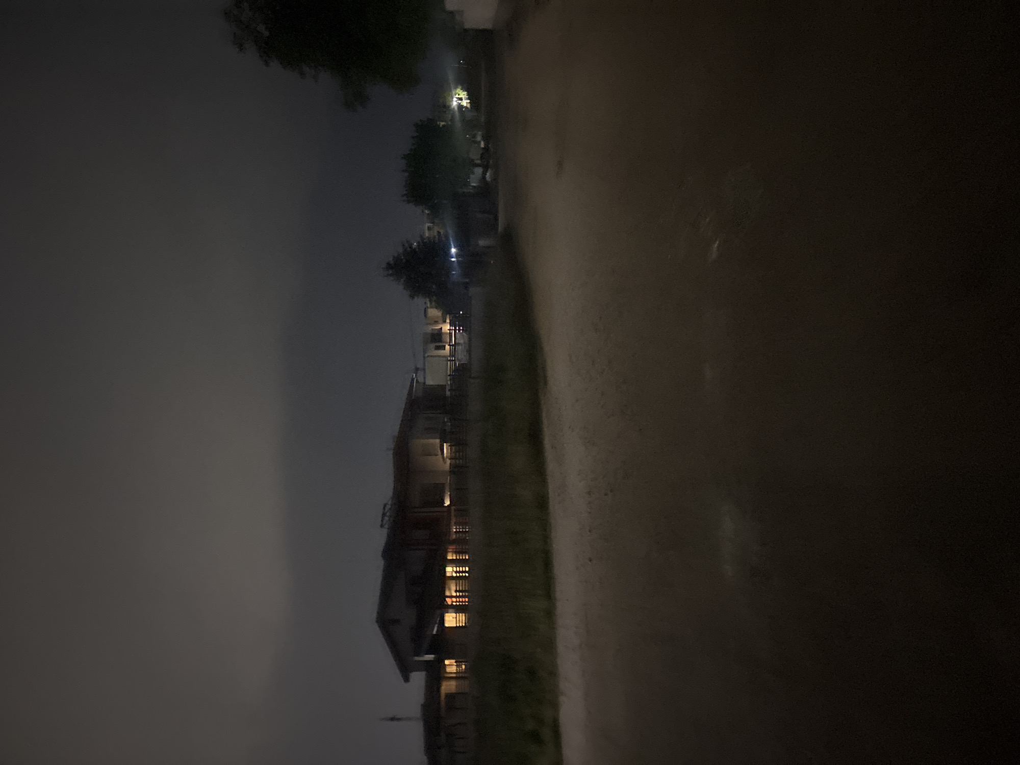 A night scene. A narrow paved road takes up the right half of the image. On the left, there is a building with lit windows, suggesting it is inhabited. Beyond the building, trees and a hazy sky are visible. Based on visual elements alone, determining a specific location within Plovdiv, Bulgaria, is not possible.
