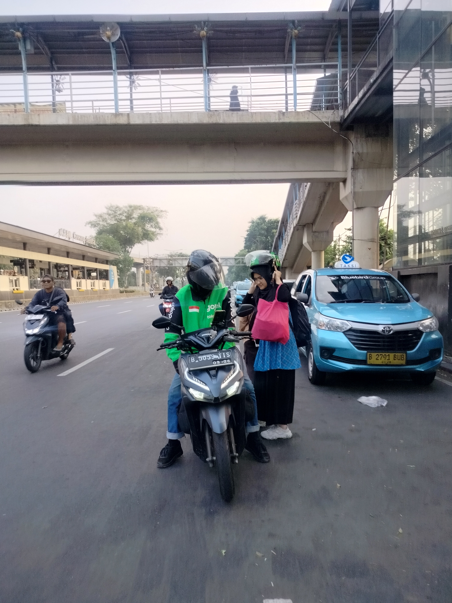 In East Jakarta, Indonesia, on a road beneath an elevated pedestrian bridge, a Gojek motorcycle rider wearing a green jacket and black helmet is positioned in the foreground. To the rider's right, a woman in a black hijab and blue skirt, carrying a pink bag, is standing. Behind them, a light blue Blue Bird taxi is stopped on the road. Further in the background, another motorcycle rider in a black jacket and helmet is visible, along with a building and trees. The sky is overcast, and the lighting suggests it is either early morning or late afternoon.