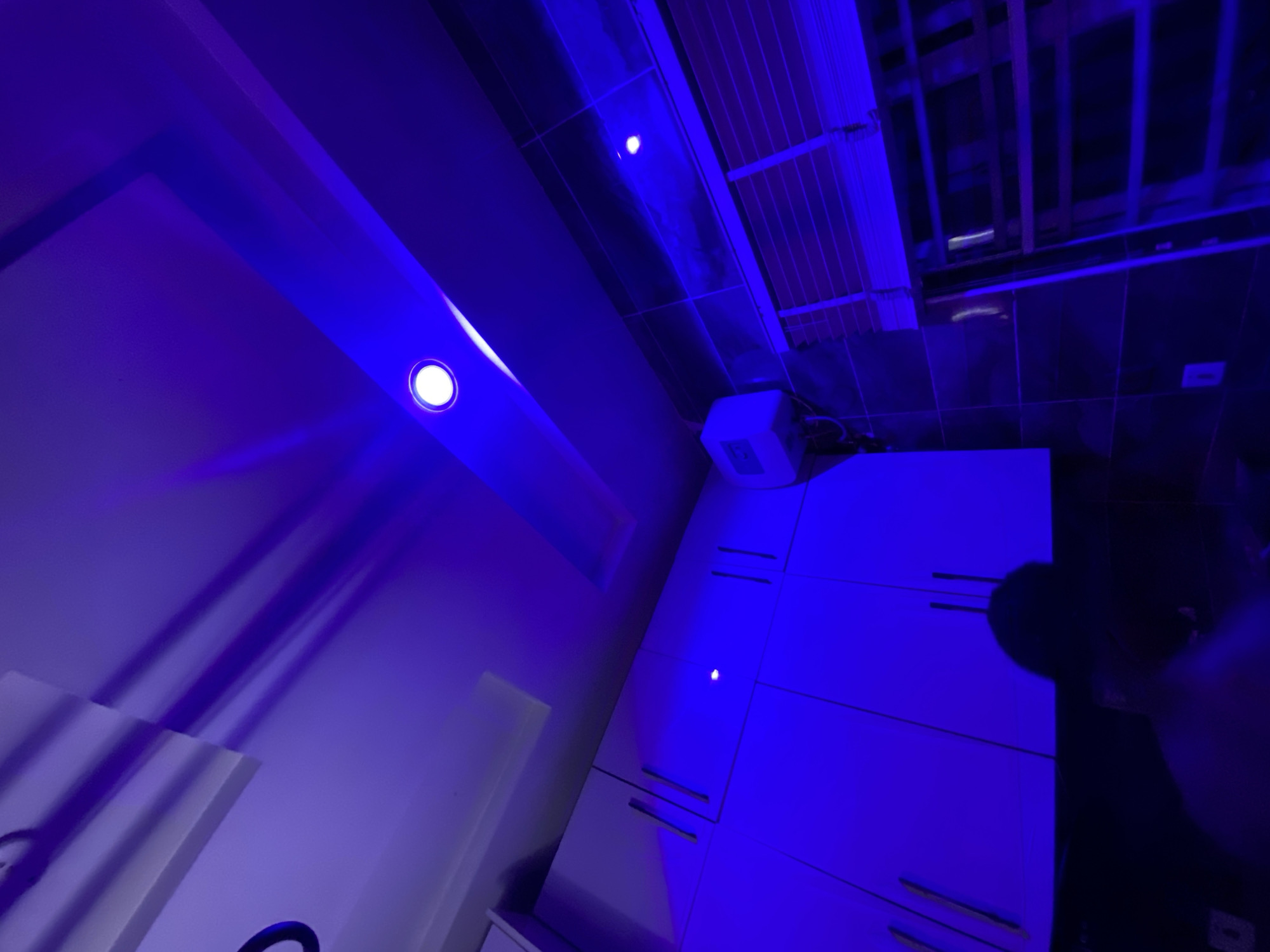 The image presents an interior view under blue lighting. Visible elements include a ceiling with exposed beams, light fixtures, a wall, and white cabinetry. The specific location within Lagos, Nigeria, is not discernible from the visual information provided. No people are fully visible, though there is a partial view of a dark human silhouette. There are no evident actions or interactions taking place in the depicted scene.