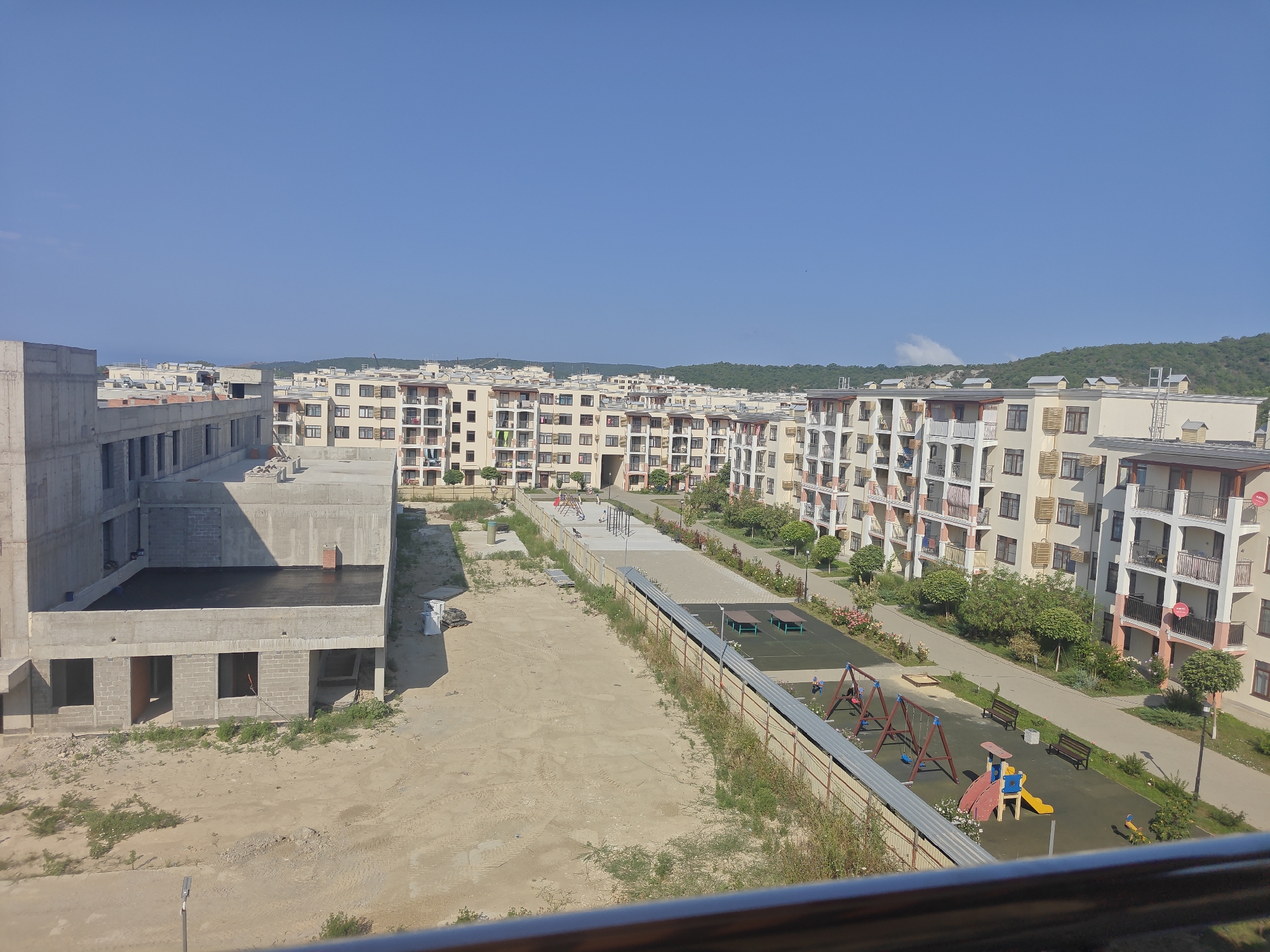 This is an aerial photograph taken in the city of Sukko, Russia, on a clear, sunny day. The image primarily depicts a residential complex under construction and a completed section with amenities.
In the foreground, a concrete building in the early stages of construction is visible. It has unfinished brickwork and large openings where windows and doors will be placed. A flat, black surface covers the top of this structure, possibly a waterproof layer. The ground around this building is sandy and uneven, with some patches of dry grass and tire tracks.
Adjacent to the construction site, a completed residential complex stretches into the background. These buildings are multi-story apartment blocks with balconies and uniformly colored facades. They are arranged in rows, creating a dense urban landscape.
Between the unfinished and completed buildings, a recreational area is present. This area includes a children's playground with swings, a slide, and a sandbox. There are also picnic tables and benches, suggesting a communal space for residents. A paved pathway runs through this area, and it is lined with trees and rose bushes.
The sky is a vibrant, cloudless blue, indicating fair weather. The overall scene captures a developing residential area, showcasing both the progress of construction and the established living spaces with their associated amenities.