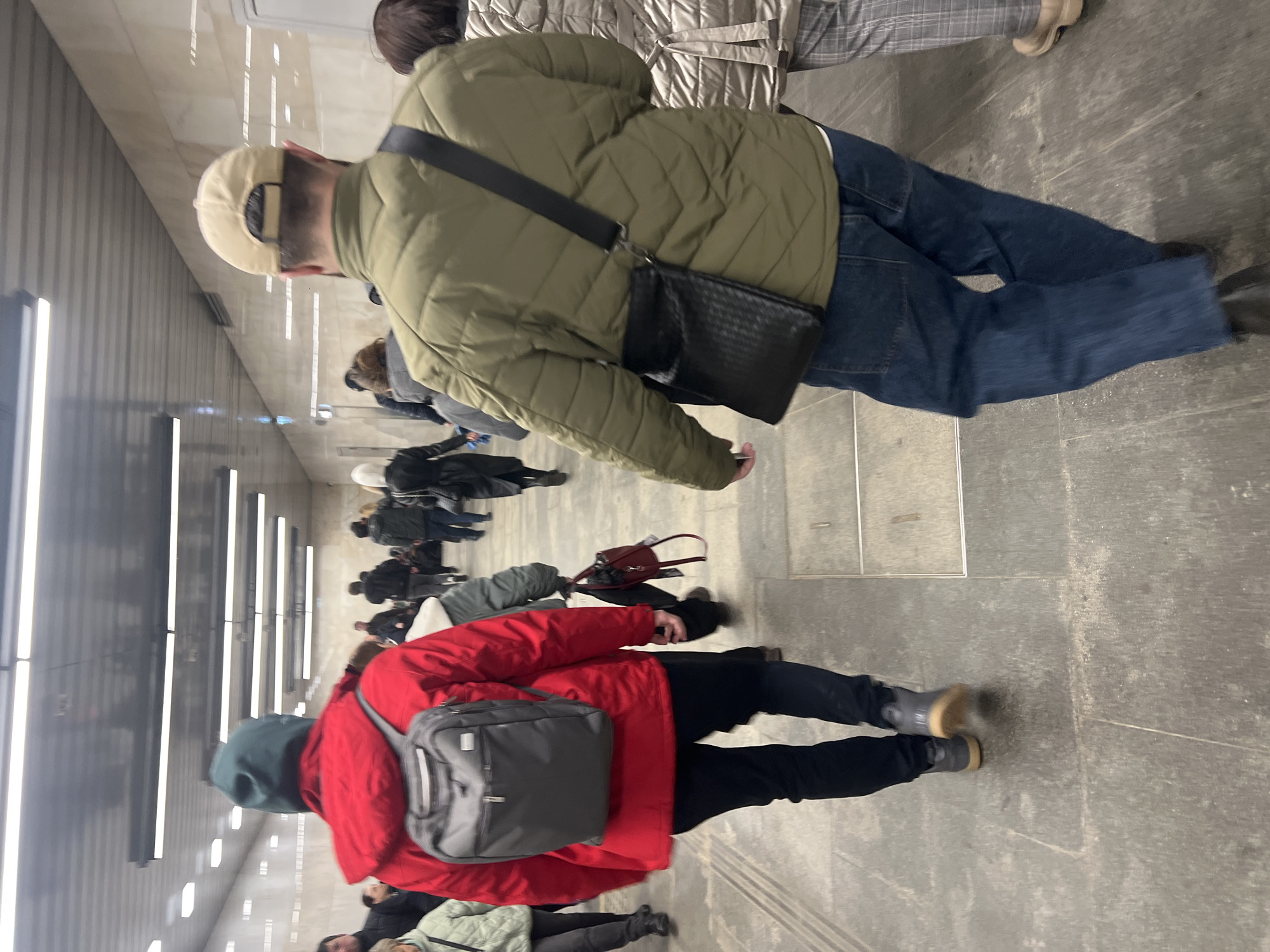 The image shows a group of people walking in a hallway with bright, linear lighting. The scene is captured from an elevated perspective. The foreground features two individuals prominently. One person wears a red jacket with a gray backpack and black pants, while the other wears a green quilted jacket, blue jeans, and a tan baseball cap. The person with the green jacket also has a black crossbody bag. The background shows more people walking further down the hallway, which appears to be a corridor in a public space. The location is likely an underground passage, possibly a subway station in Khimki, Russia.