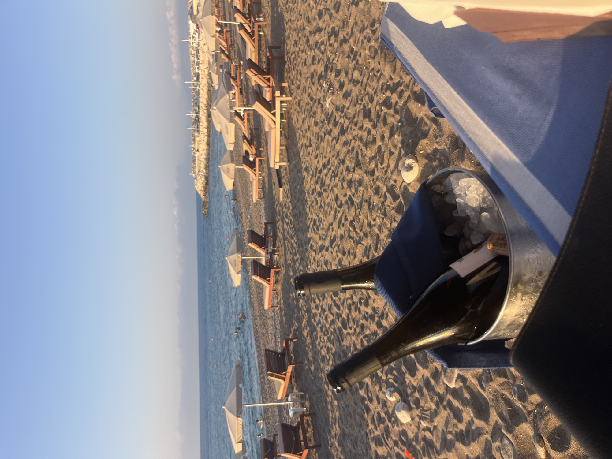 A sandy beach in Polis Chrysochous, Cyprus, is shown at sunset. Two bottles of white wine are in a bucket of ice in the foreground, placed on a blue towel on the sand. In the background, lounge chairs and umbrellas are set up along the beach, with the calm sea and a breakwater visible in the distance. The sky is a clear, pale blue, with soft clouds near the horizon. A few people are in the water.