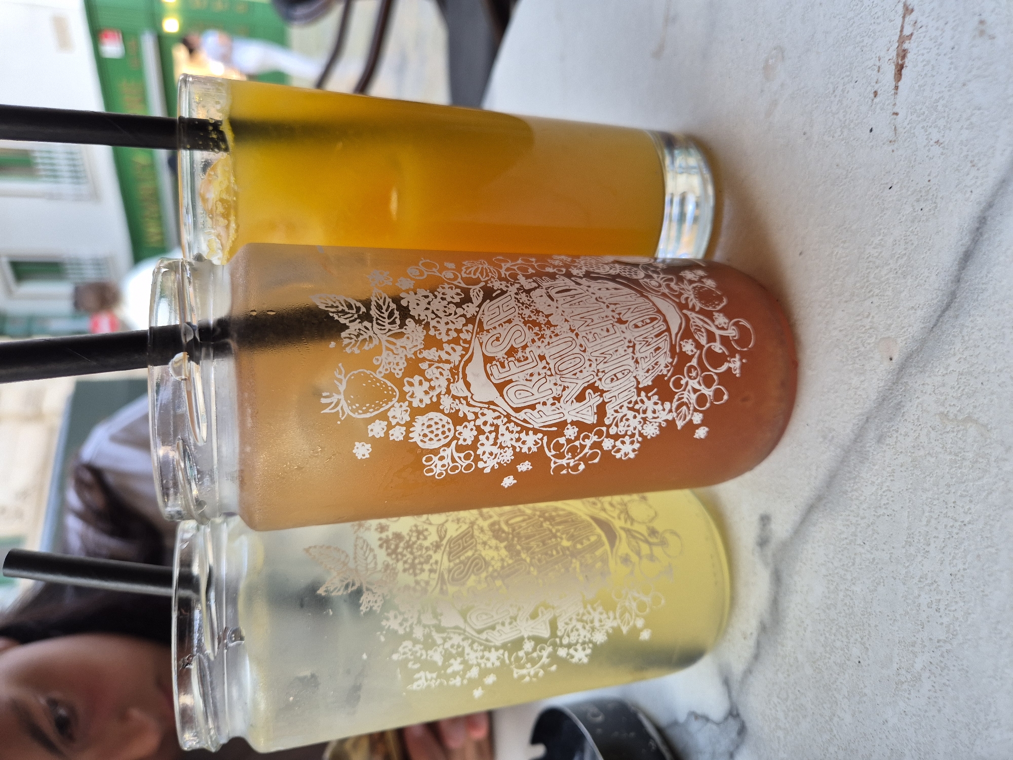 Three glasses of iced beverages with straws are arranged on a marble-patterned table. The glass on the left contains a pale yellow liquid and has a white, floral decal. The middle glass holds an orange-colored drink with a similar white, floral decal featuring a strawberry illustration. The rightmost glass contains a vibrant orange beverage. In the background, blurry figures are visible, with a building facade that suggests an outdoor cafe setting in Valletta, Malta. The overall scene captures a casual moment, likely at a cafe or restaurant.