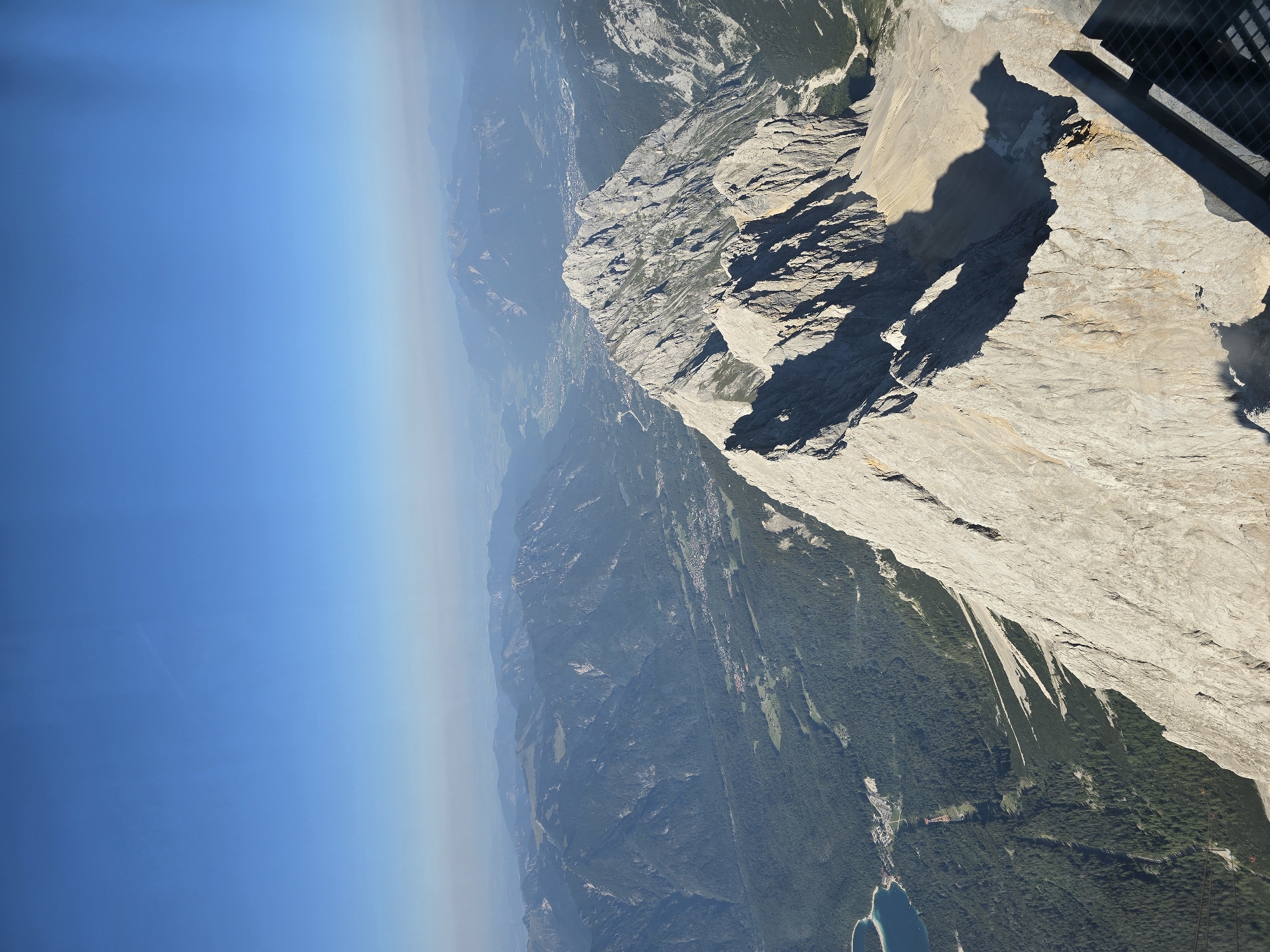 The image captures a high-altitude panoramic view of the Zugspitze region near Grainau, Germany. The foreground features the rugged, rocky terrain of a mountain peak, casting long shadows in the afternoon sun. In the mid-ground, a sharp mountain ridge dominates the view, with steep slopes and jagged peaks. Further in the distance, a valley unfolds, dotted with a small town or village, characterized by buildings with red roofs, nestled among dark green coniferous forests. A bright blue lake is visible on the left side, adjacent to the tree line. The background showcases a vast expanse of rolling mountains stretching towards a hazy horizon under a clear blue sky. A metal railing and a grated platform are partially visible in the bottom right corner, suggesting a viewpoint or observation deck. No people are discernible in the image.
