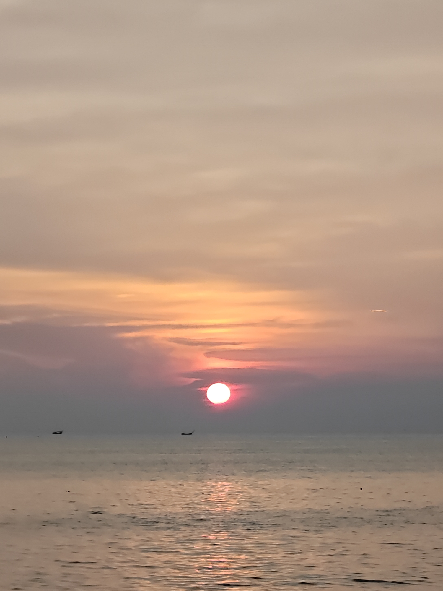 The image shows a sunset over the ocean. The sun is a bright red orb near the horizon, reflecting off the water below. There are two small boats silhouetted against the light on the horizon. The sky is a mix of orange, pink, and gray clouds. Based on the prompt, the location is presumed to be in or near Lhokseumawe, Indonesia.