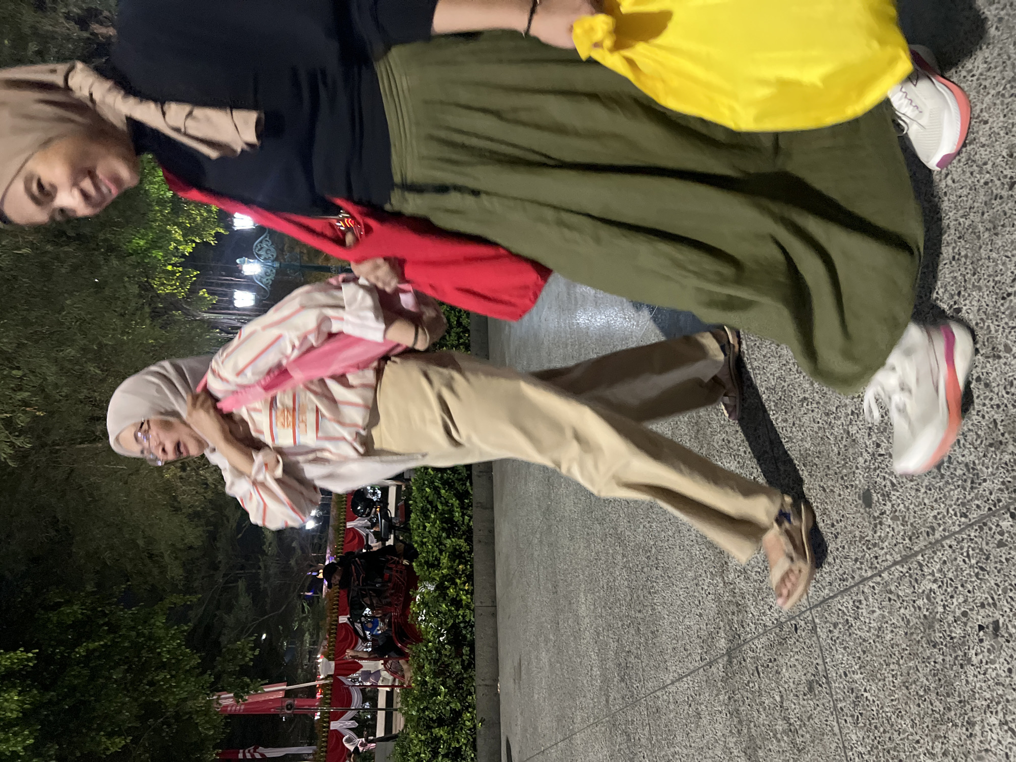Two women are walking on a paved surface in front of a decorative display at night. The woman on the left wears a light-colored hijab, glasses, and a striped long-sleeved shirt with tan pants and sandals. She is carrying a pink bag. The woman on the right wears a tan hijab, a black long-sleeved shirt, and a long olive green skirt. She is carrying a red bag and a yellow bag. The background includes trees, streetlights, and a display with red and white banners, suggesting a festive or event-related setting, likely in the city of Yogyakarta, Indonesia. The lighting suggests it is after sunset.