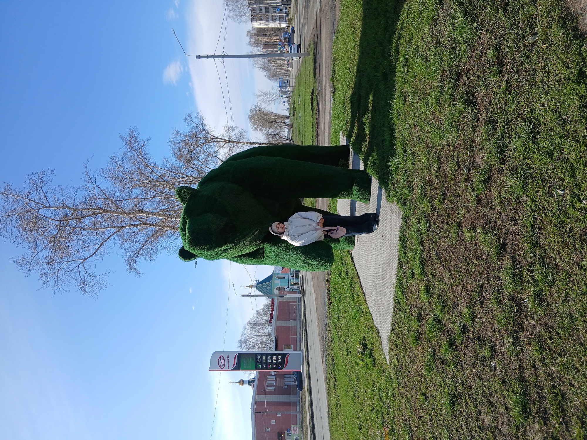 A person lies atop a green, moss-covered sculpture of a bear, located on a grassy area next to a sidewalk and road in the city of Unknown, Unknown. The person is wearing a white jacket, dark pants, and a white hat. Behind the sculpture and person, a street with buildings, a gas station price sign, and a church are visible. A bare tree and blue sky fill the upper portion of the frame.