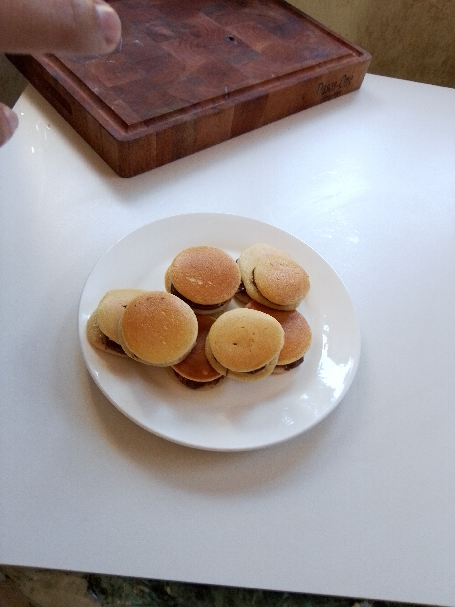 A plate of mini pancakes is on a white table, next to a wooden cutting board. The cutting board has a logo that reads "Pasco-One". A hand is partially visible in the upper left corner of the image. The scene appears to be indoors, possibly in a kitchen or dining area.