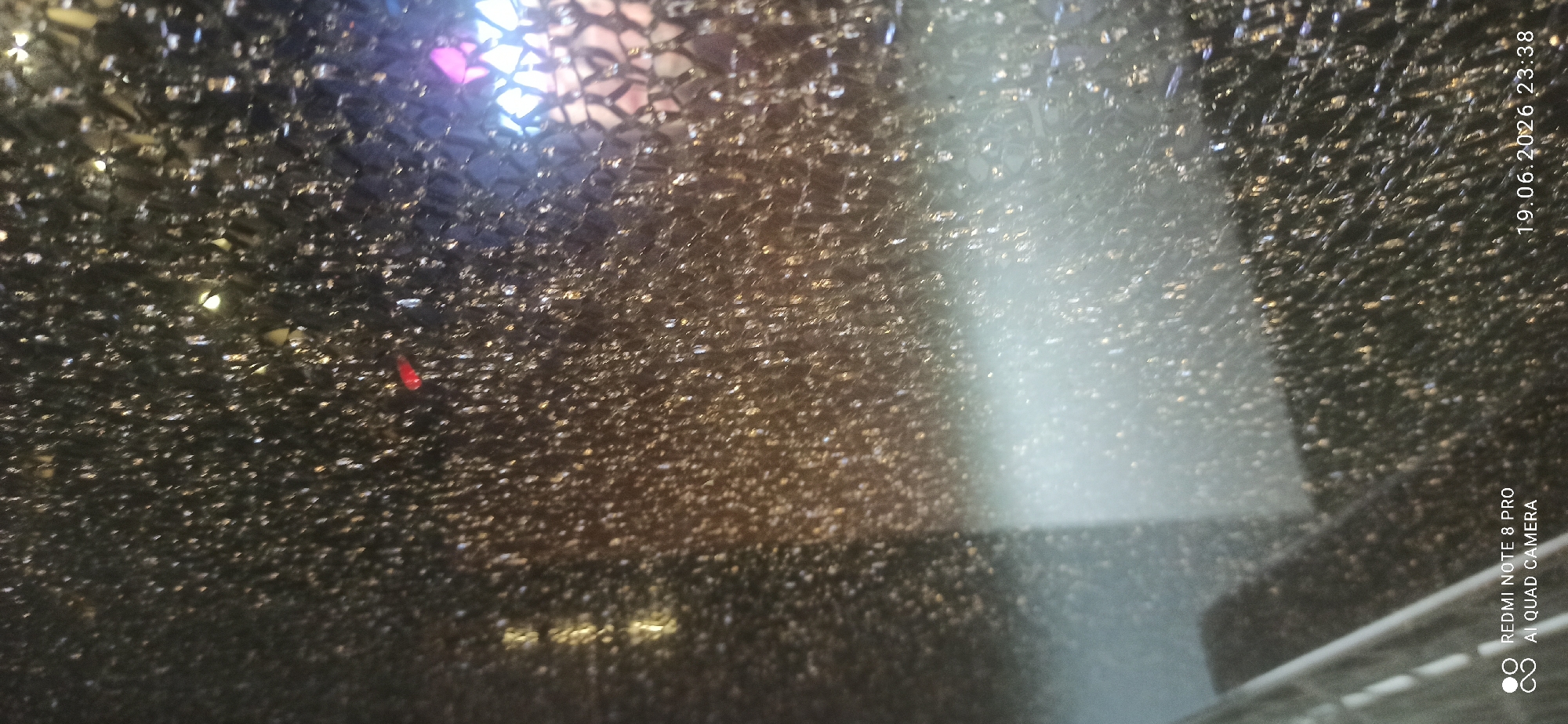 The image displays a close-up of shattered glass, likely from a window or a mirror. The glass is fractured into numerous small pieces, creating a mosaic-like pattern. The background appears dark and blurry, with some indistinct lights suggesting an indoor or nighttime setting. There is a faint, bright light source emanating from the right side of the image, and a reddish object is visible on the upper left portion of the shattered glass. Text indicating "REDMI NOTE 8 PRO AI QUAD CAMERA" and a date and time stamp "19.06.2026 23:38" are present in the lower corners of the image, suggesting it was taken by a smartphone. The overall impression is one of destruction or damage.